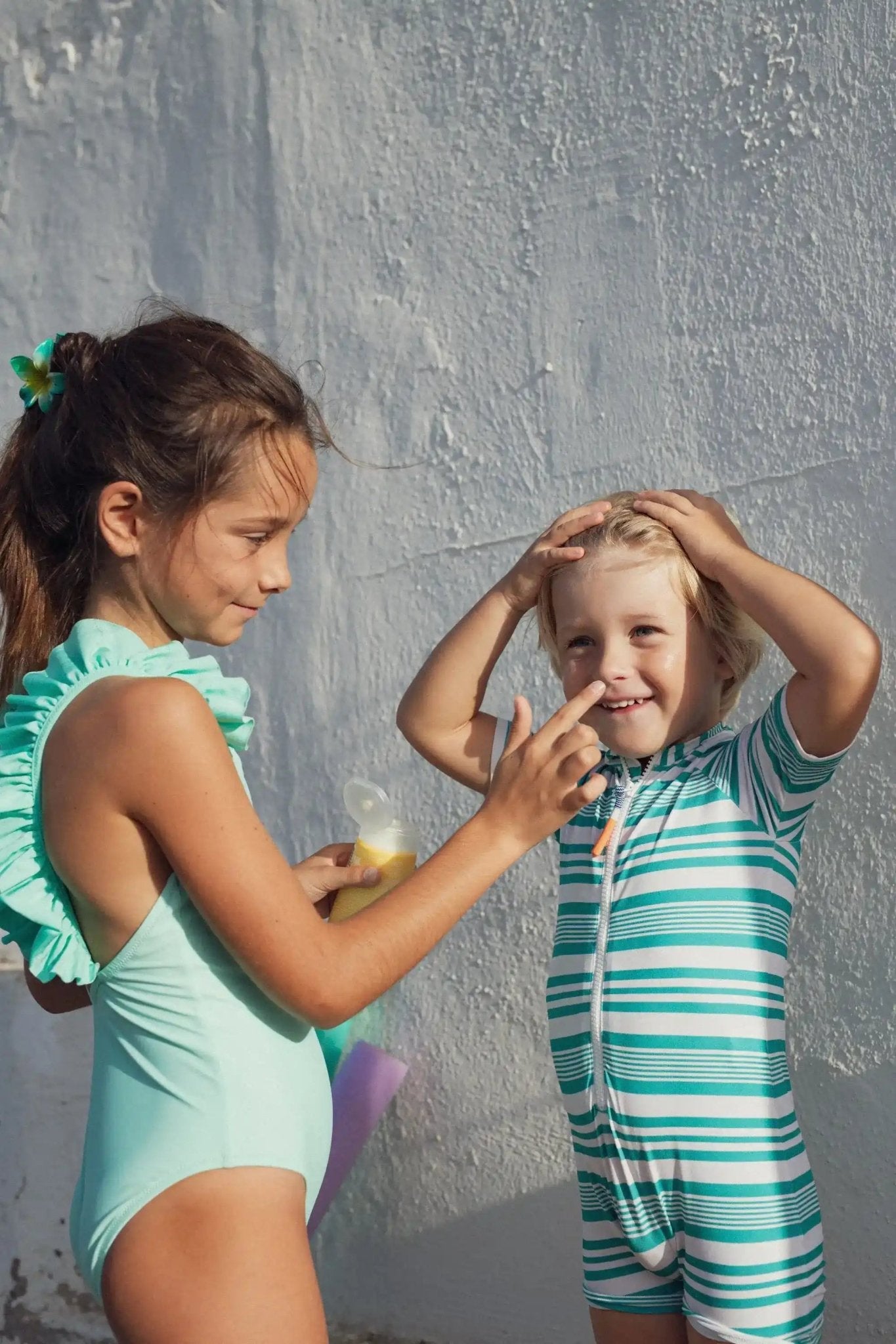 Girl applying sunscreen to boy in green and white striped short-sleeve UV swimsuit outdoors