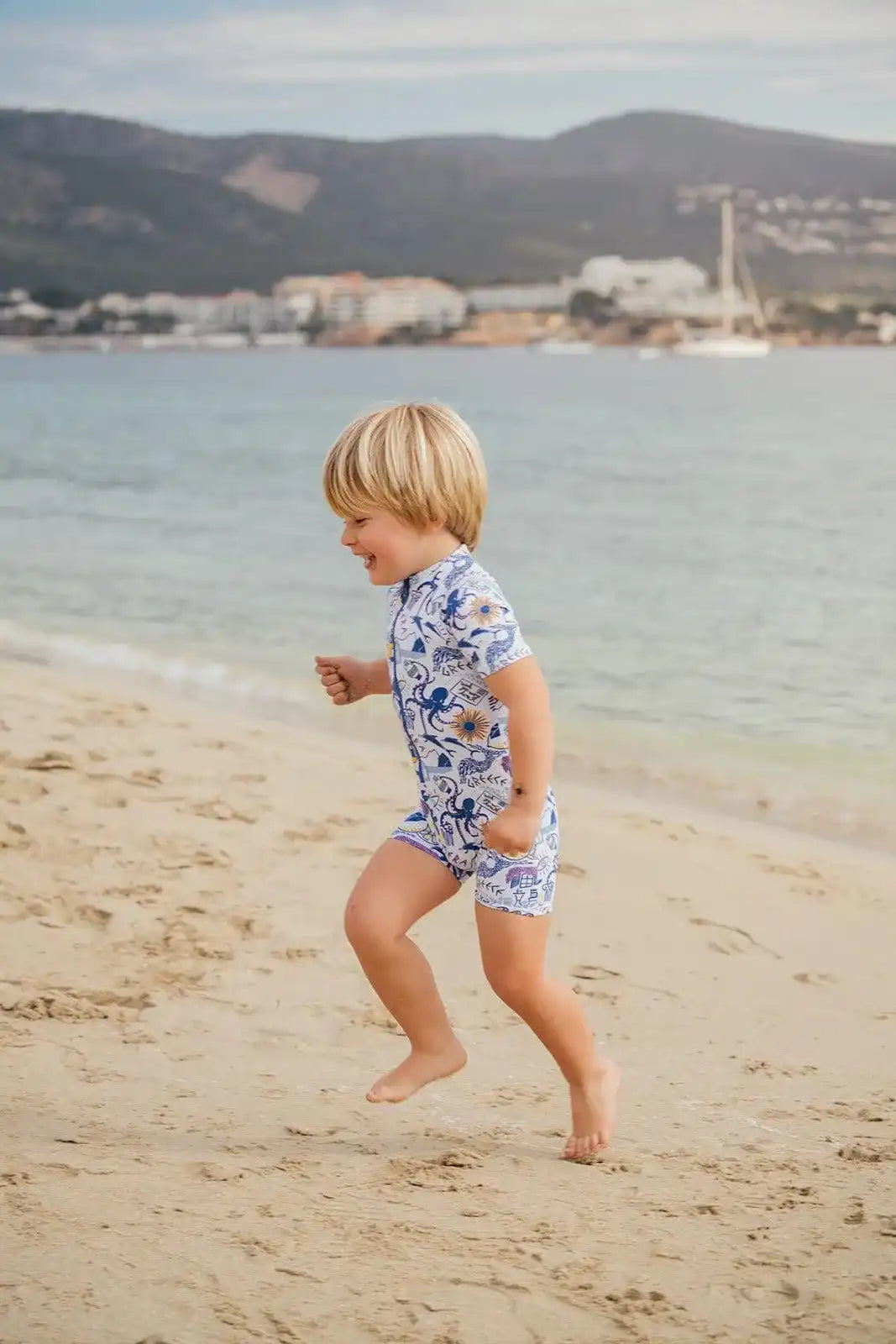 Young boy in Lison Paris anti-UV Greek motif swimsuit running on sandy beach by the sea