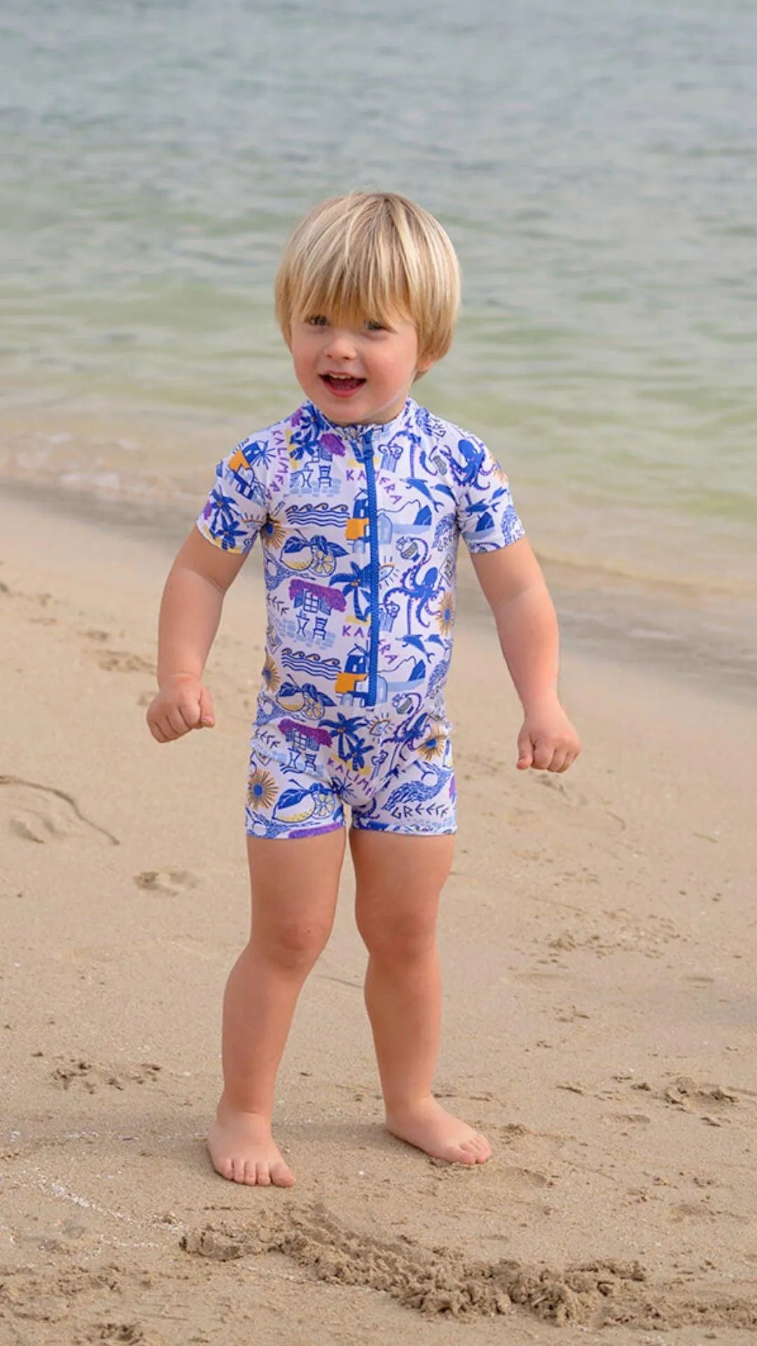 Toddler boy in short-sleeve UV protective swimsuit with blue Greek patterns standing on sandy beach