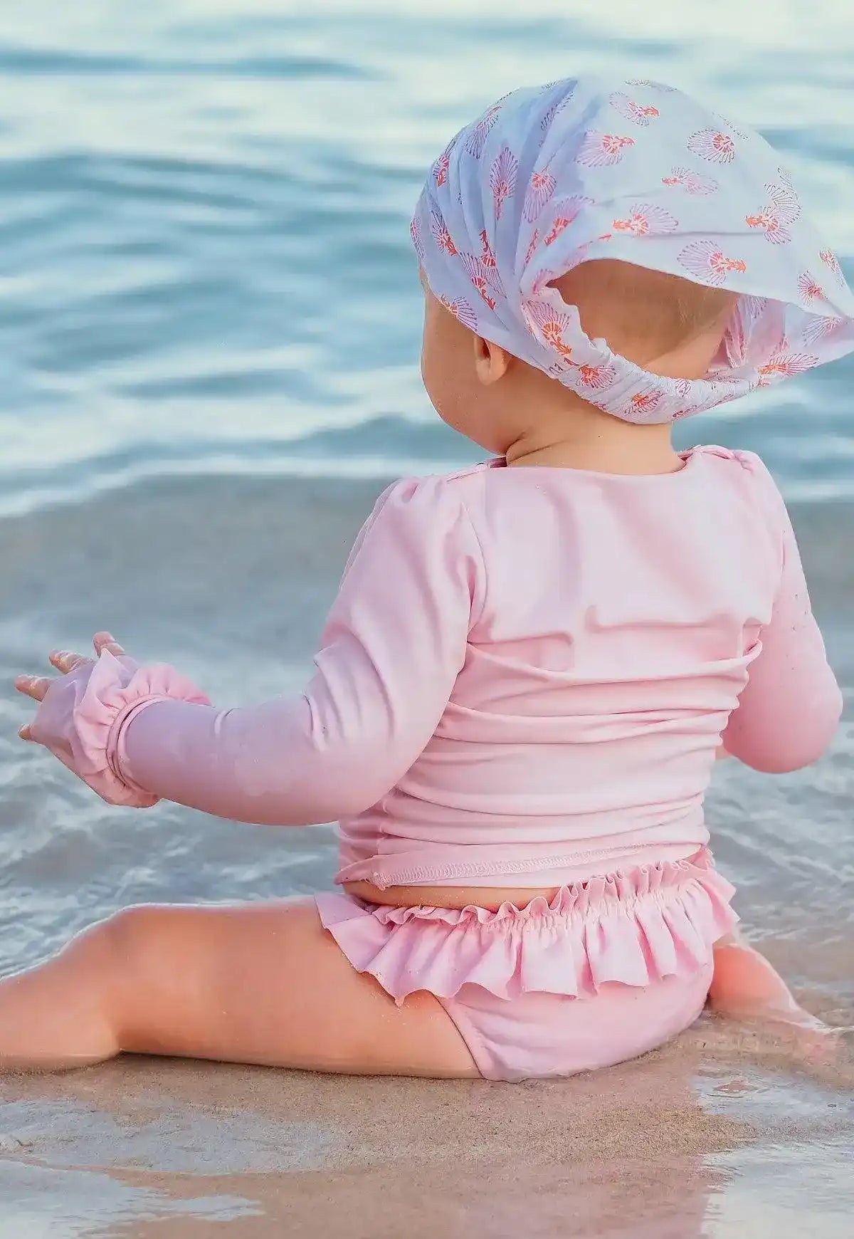 Baby girl in pale pink UV-protection swim bottoms and long-sleeve top sitting by the sea