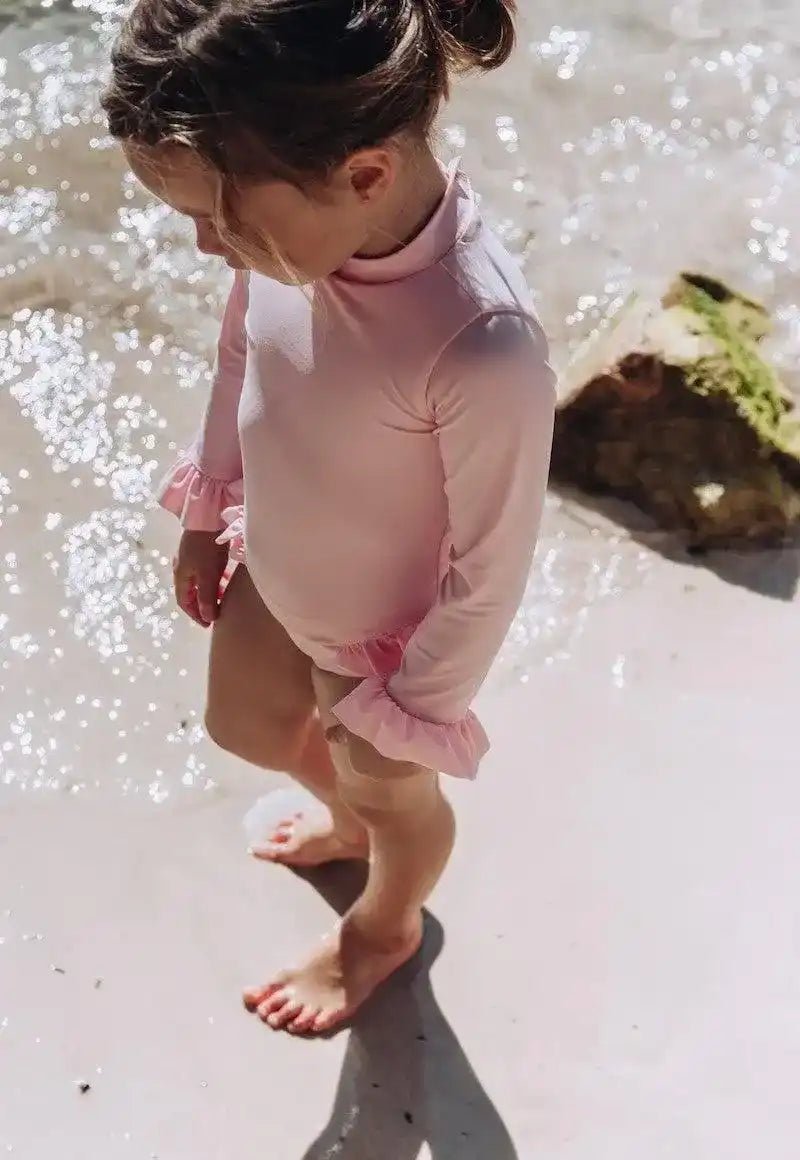 Toddler wearing pink long-sleeve UV swimsuit standing on sandy beach by the water
