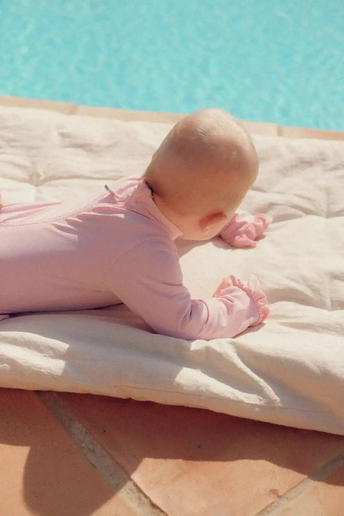 Baby in pale pink long-sleeve UV swimsuit lying by poolside on a light towel