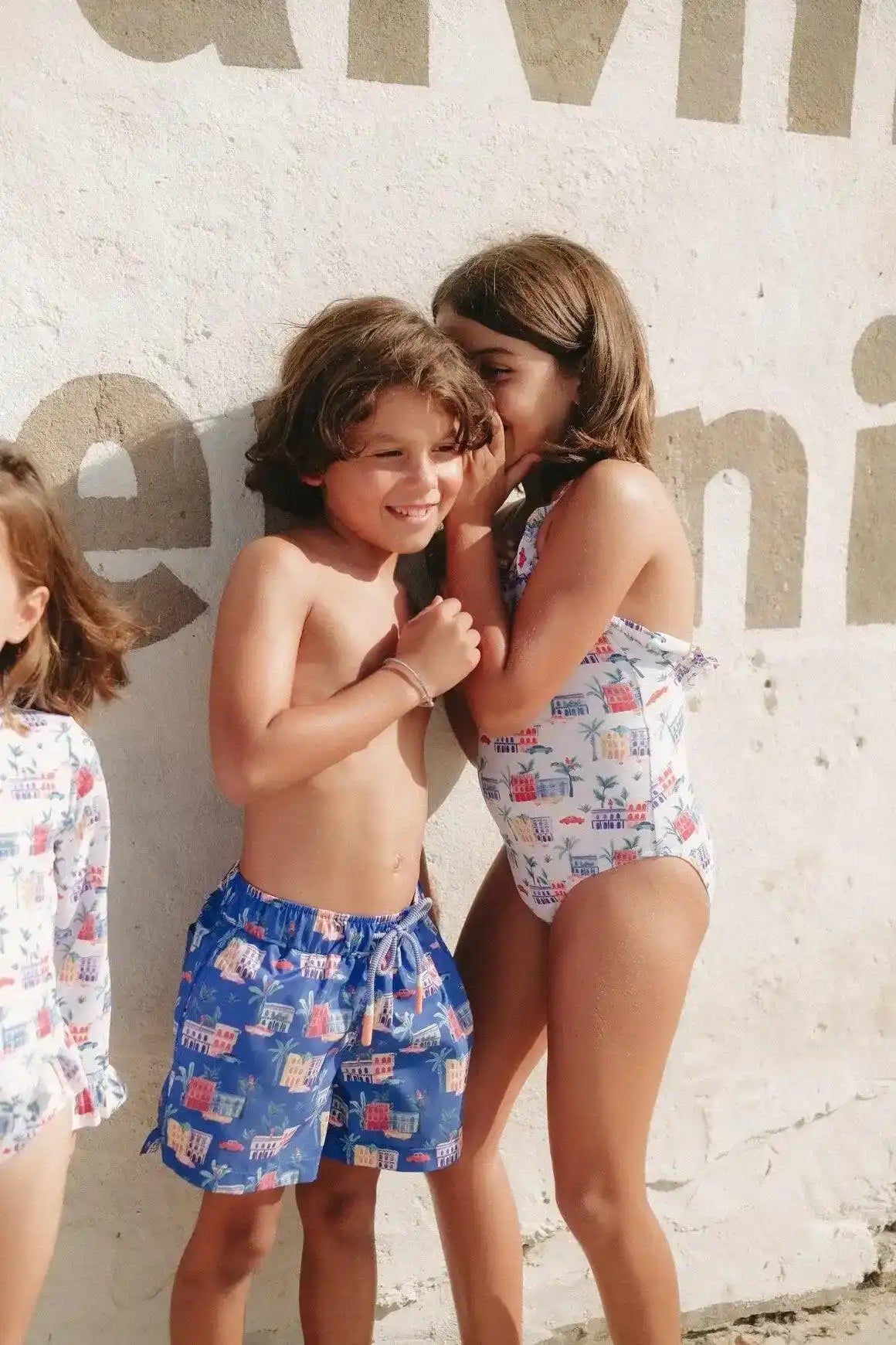 Teen boy in eco-friendly blue swim trunks and girl in printed swimsuit smiling by a wall outdoors
