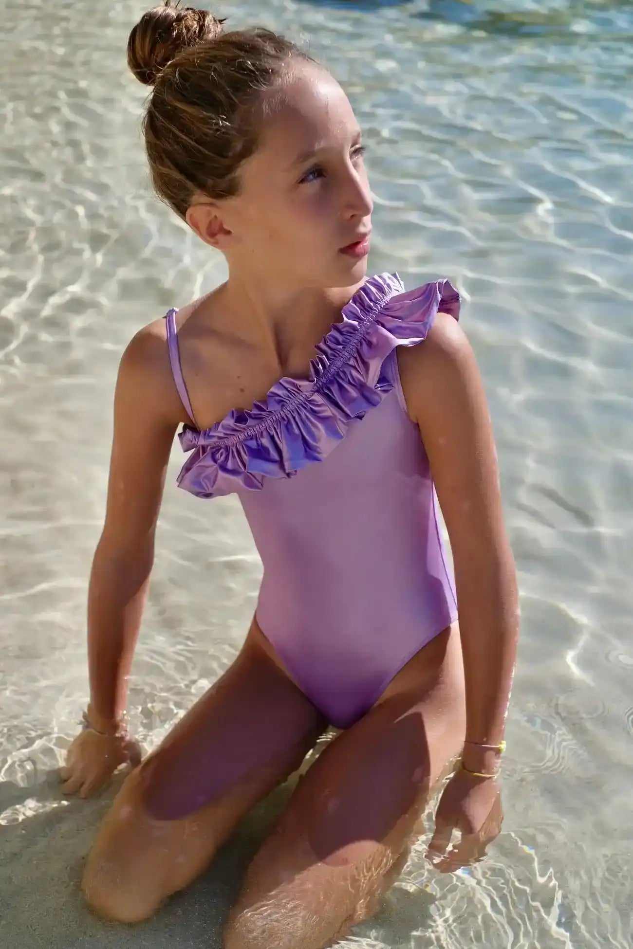 Girl in iridescent lilac asymmetric ruffle swimsuit kneeling in clear beach water