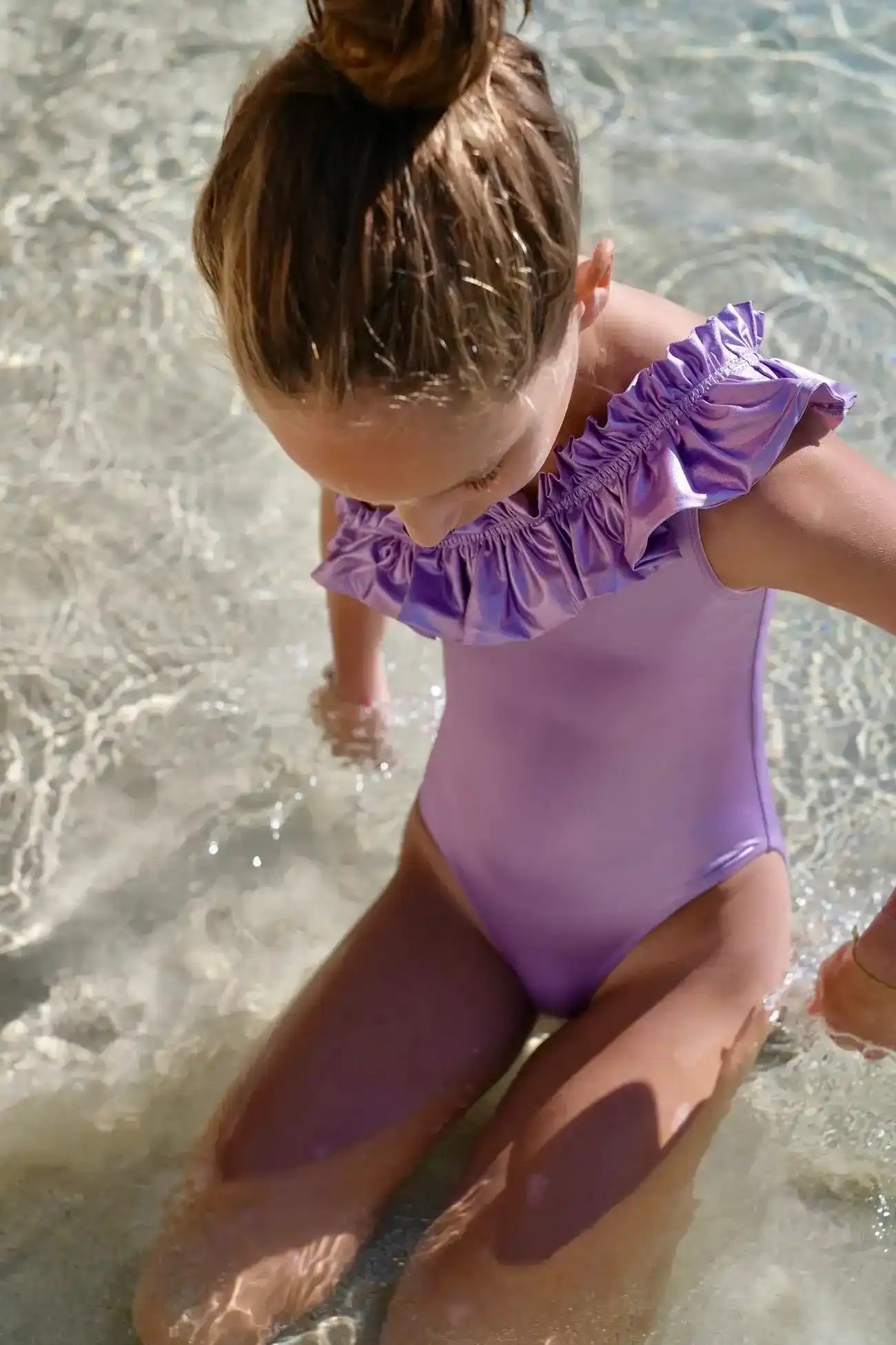 Girl wearing lilac iridescent asymmetric ruffle swimsuit kneeling in clear shallow water