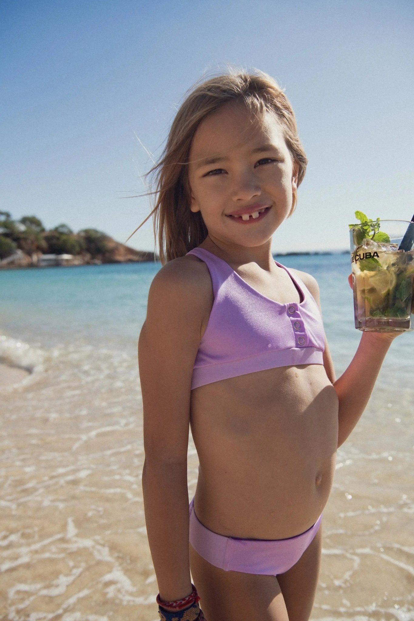 Girl in lilac two-piece swimsuit with UV protection standing on a sunny beach