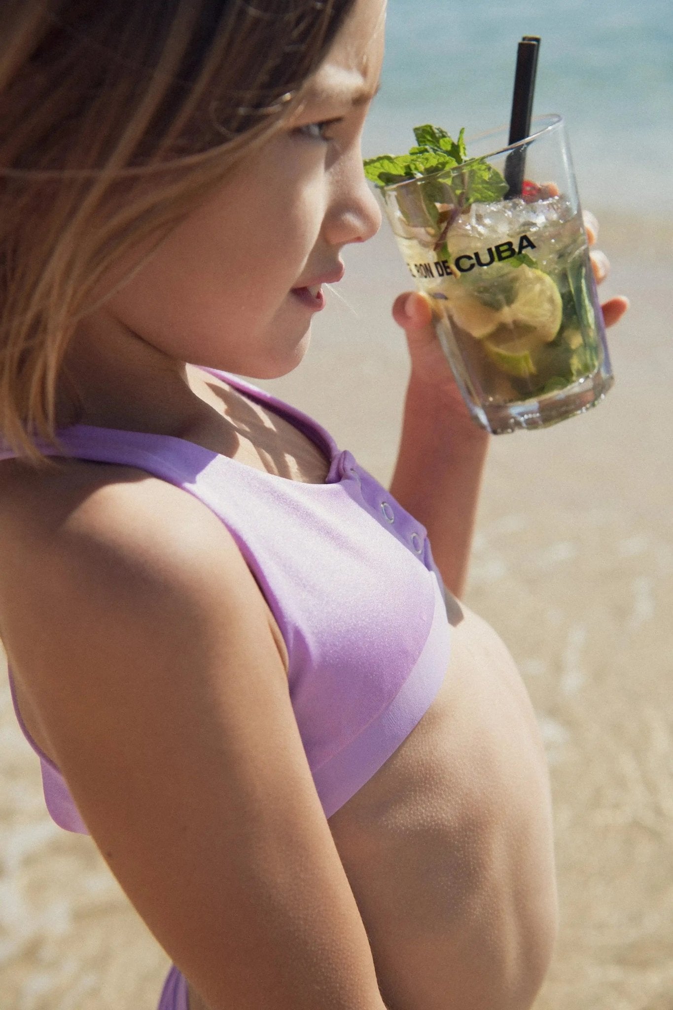 Girl in lilac two-piece UV swimwear at the beach holding a drink