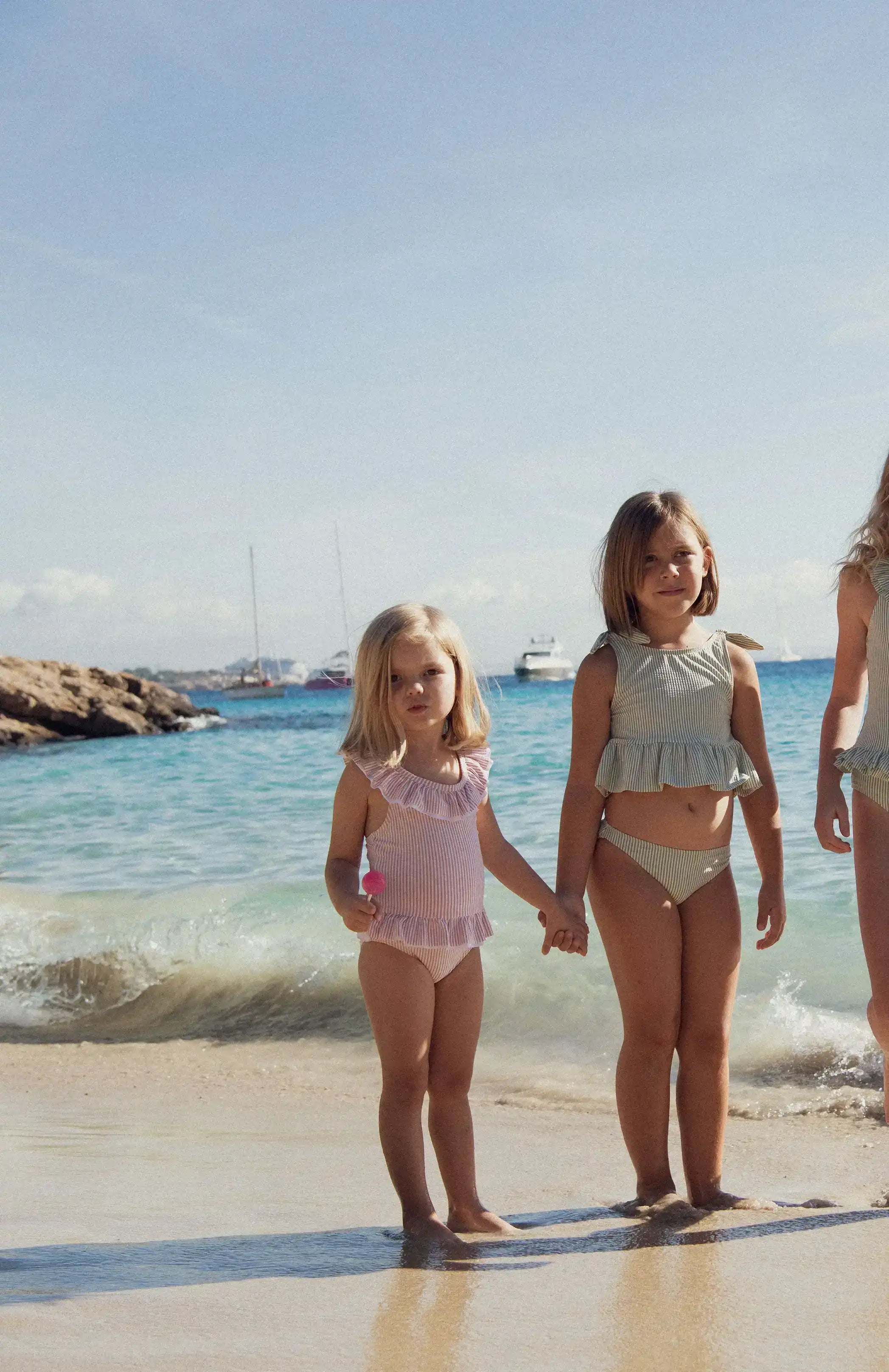 Two girls in striped ruffle swimsuits standing on a sunny beach by the sea