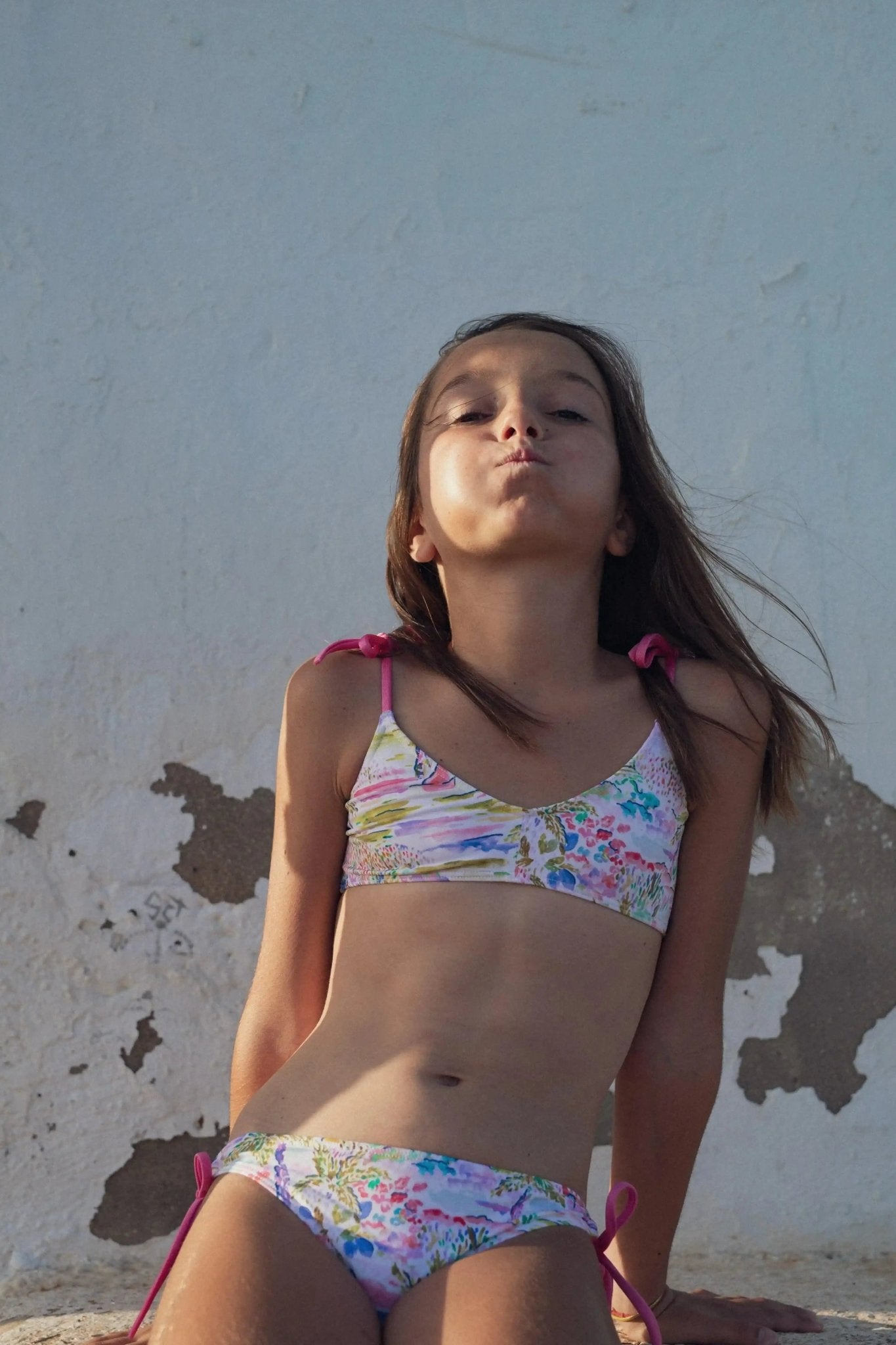 Girl in two-piece pink floral swimsuit outdoors against a sunlit textured wall