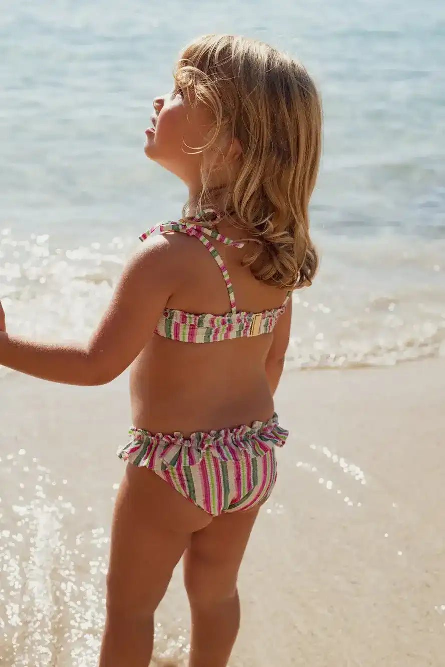 Young girl in a pink and green striped two-piece swimsuit standing on the beach by the sea
