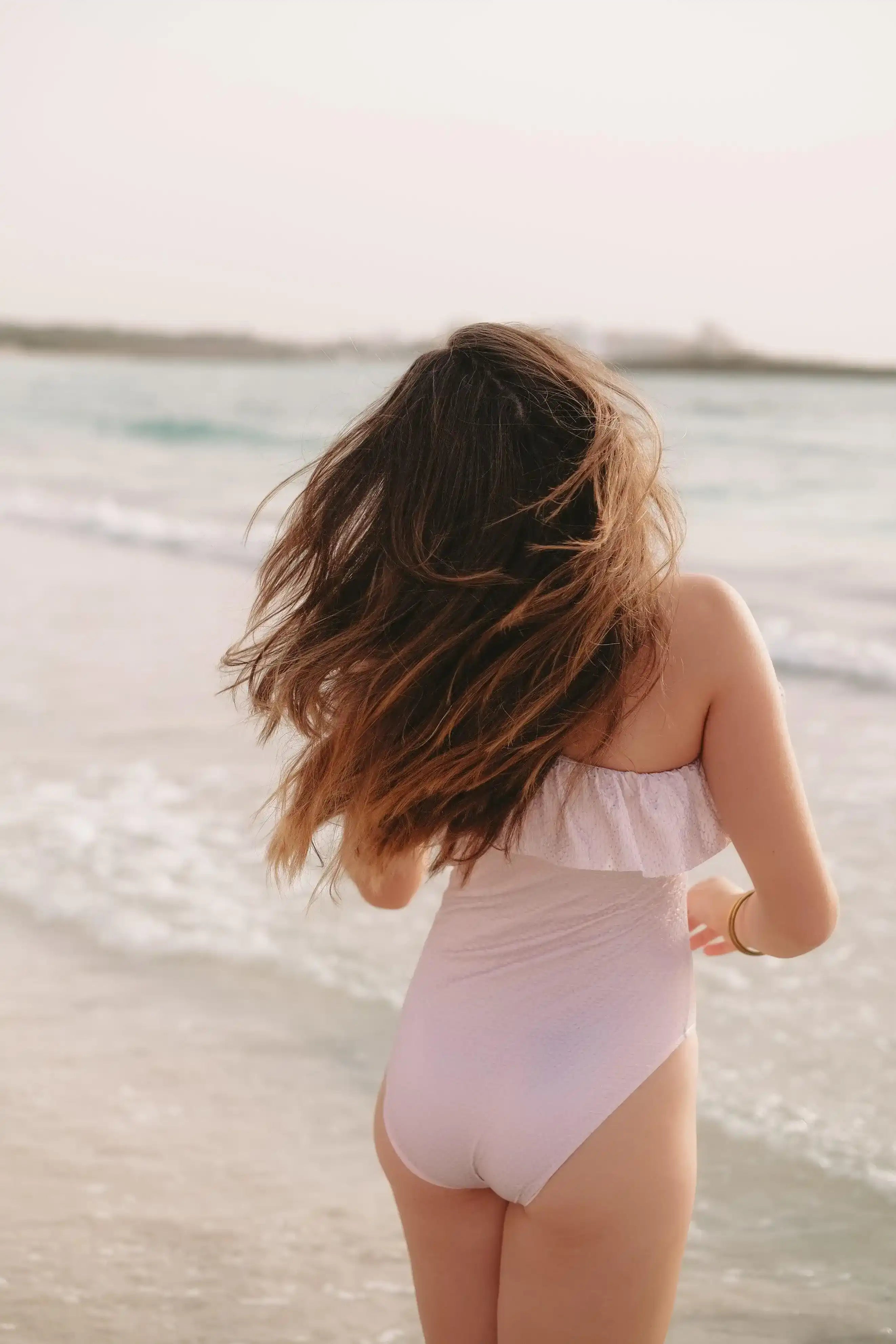 Girl in light pink asymmetric one-piece swimsuit on the beach facing the sea