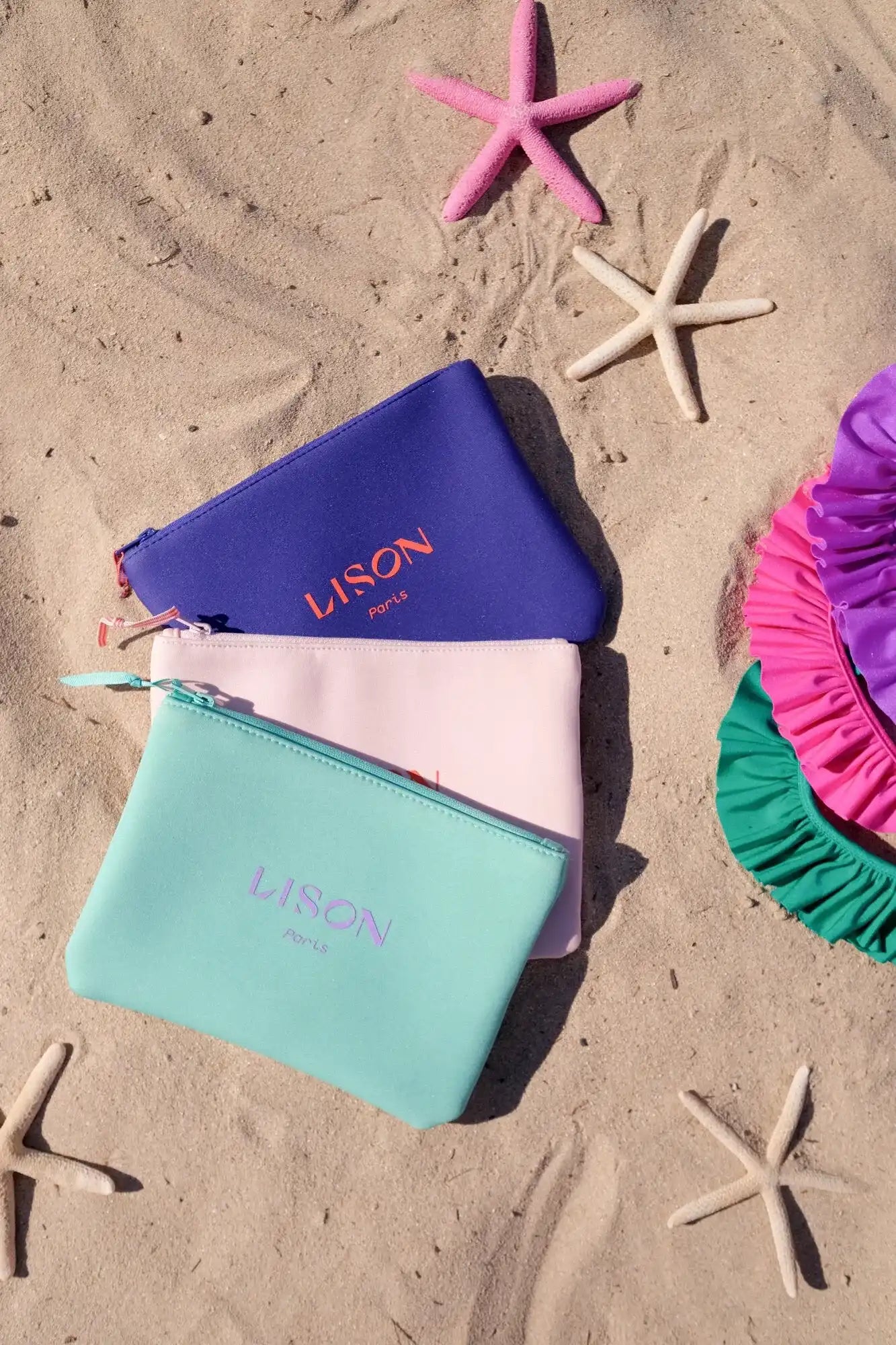 Lison Paris colorful pouches on sand with starfish and ruffled swimwear, beach scene