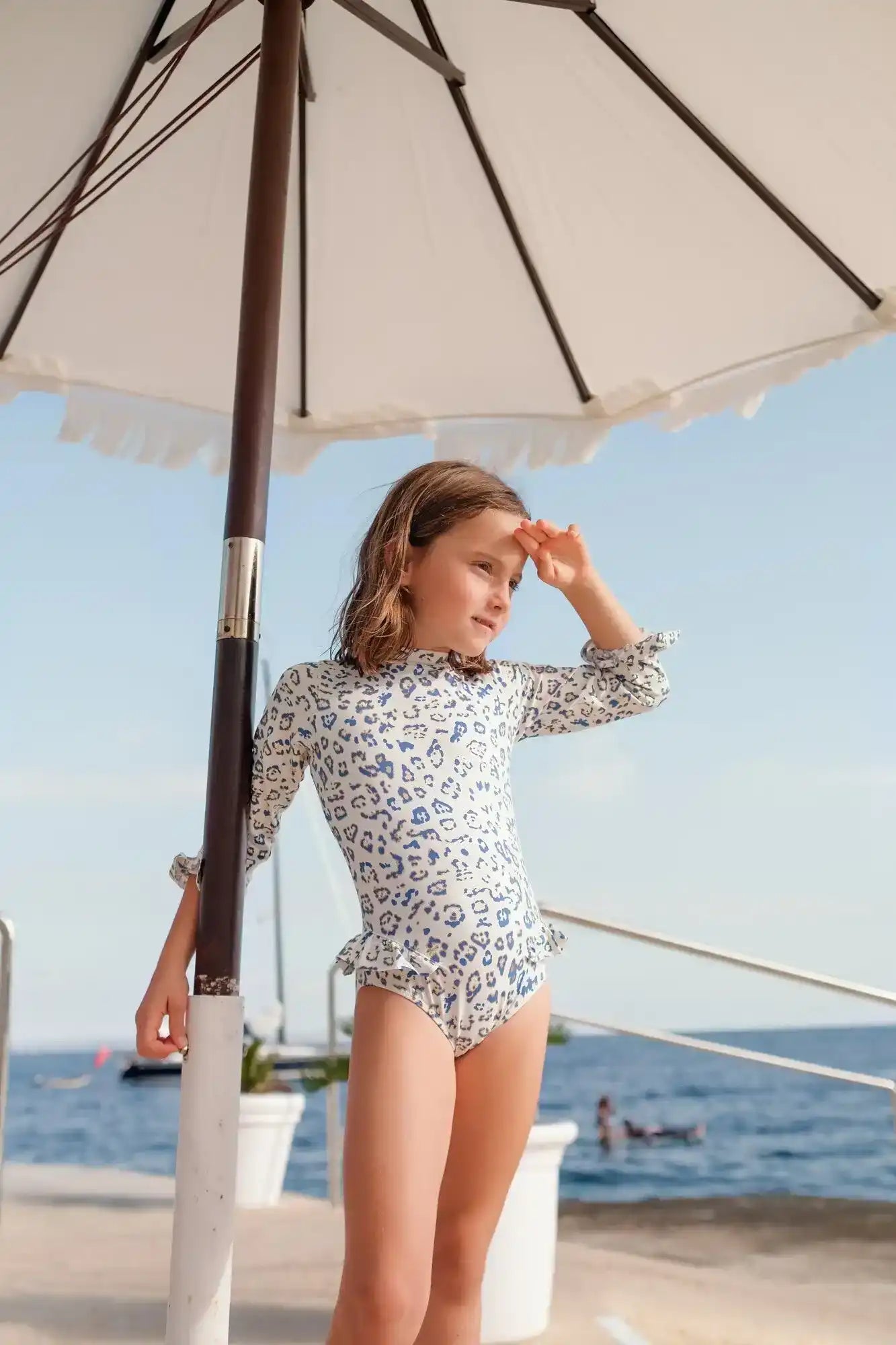 Girl wearing long-sleeve blue leopard swimsuit on beach under umbrella by the sea