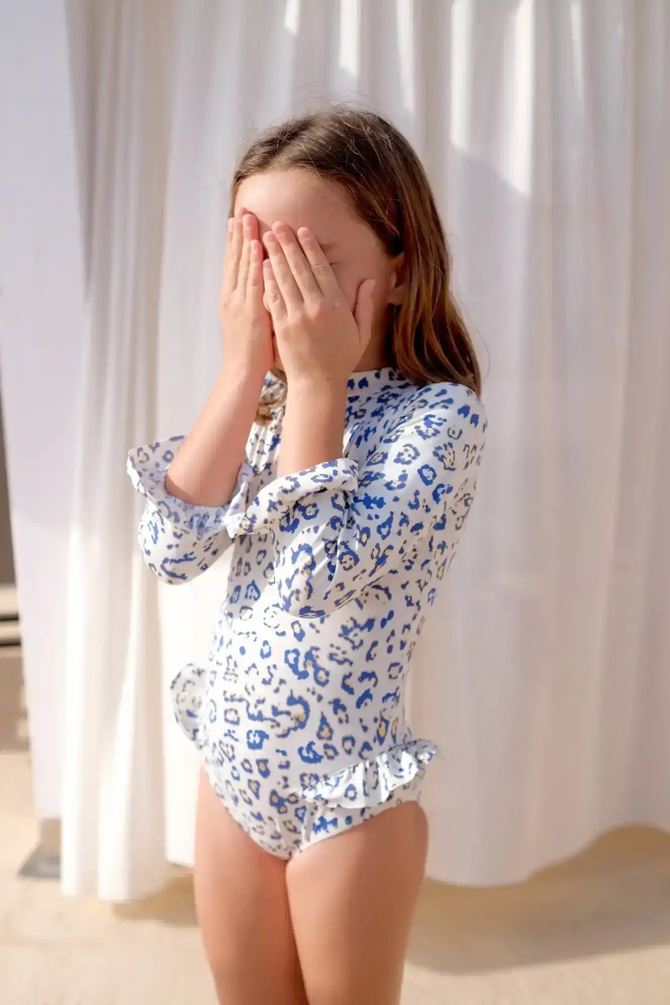 Girl in long sleeve blue leopard print baby swimsuit with ruffles, standing in sunlight
