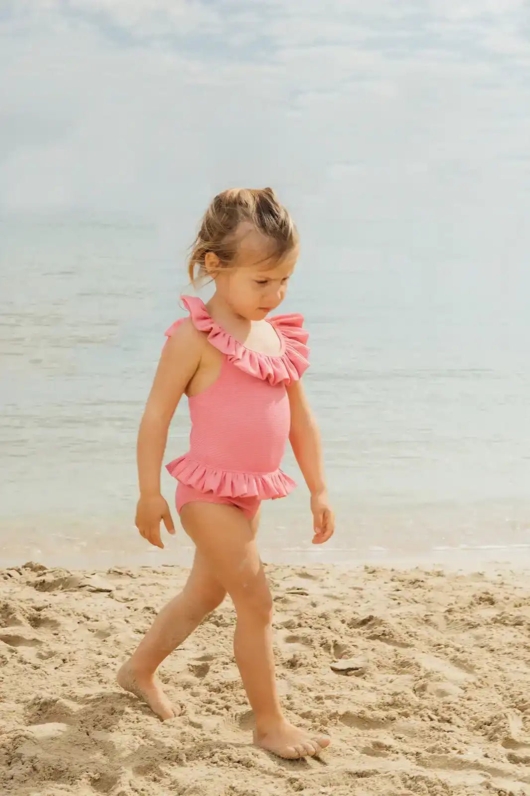 Toddler girl in pink ruffled one-piece swimsuit walking on sandy beach by the sea