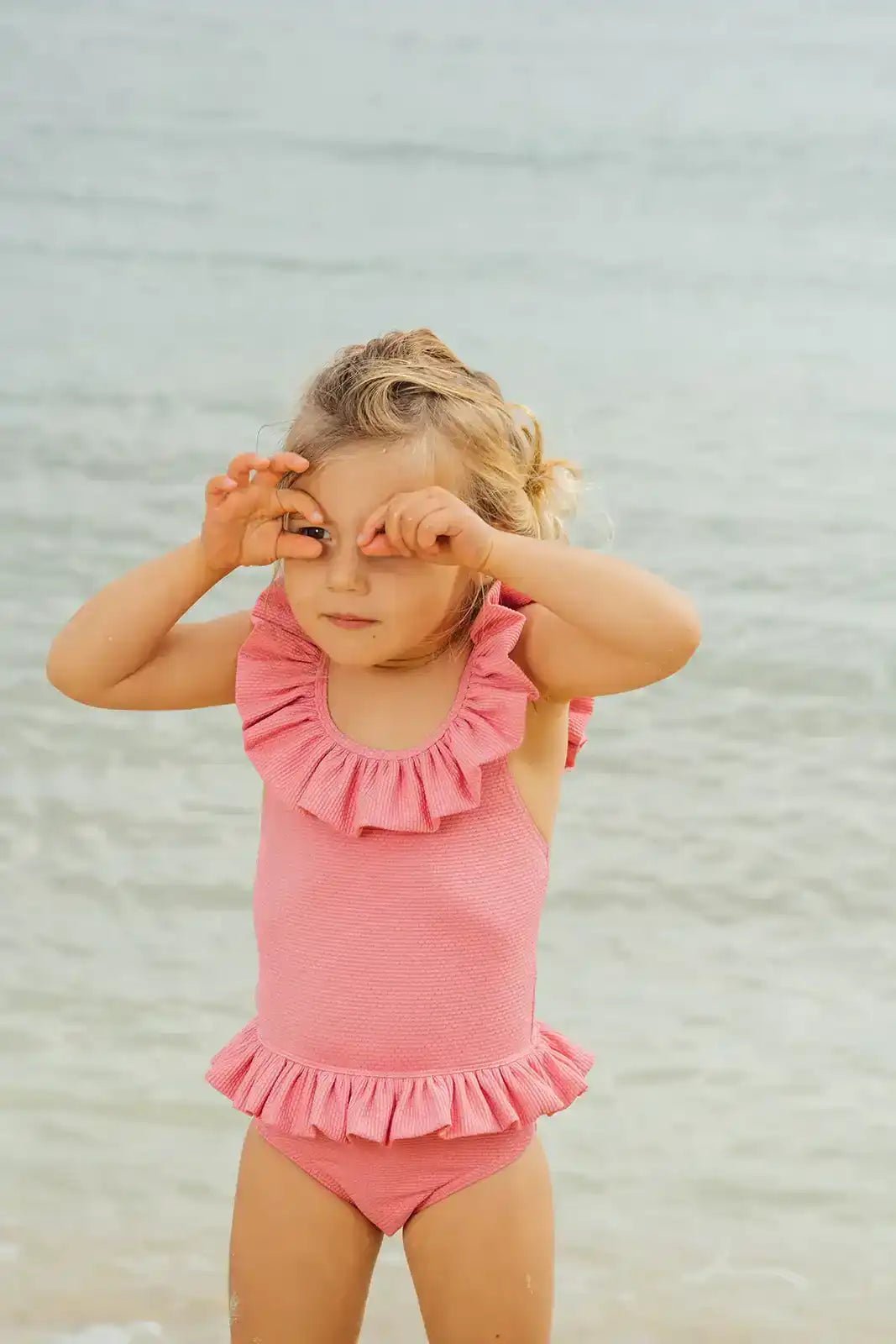 Baby girl in pink one-piece ruffle swimsuit standing on the beach by the sea