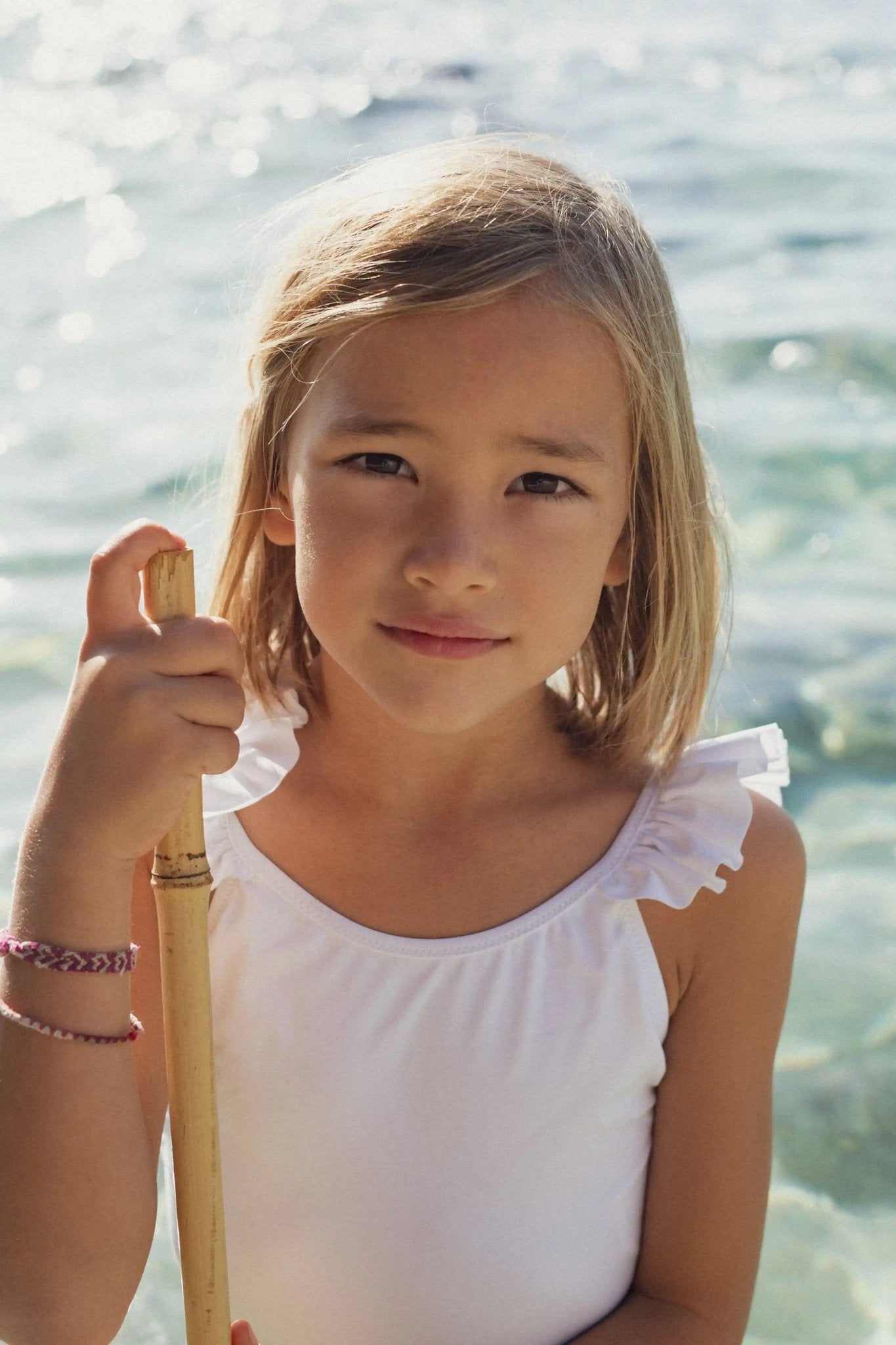 Girl wearing a white one-piece swimsuit with ruffles by the sea, holding a stick