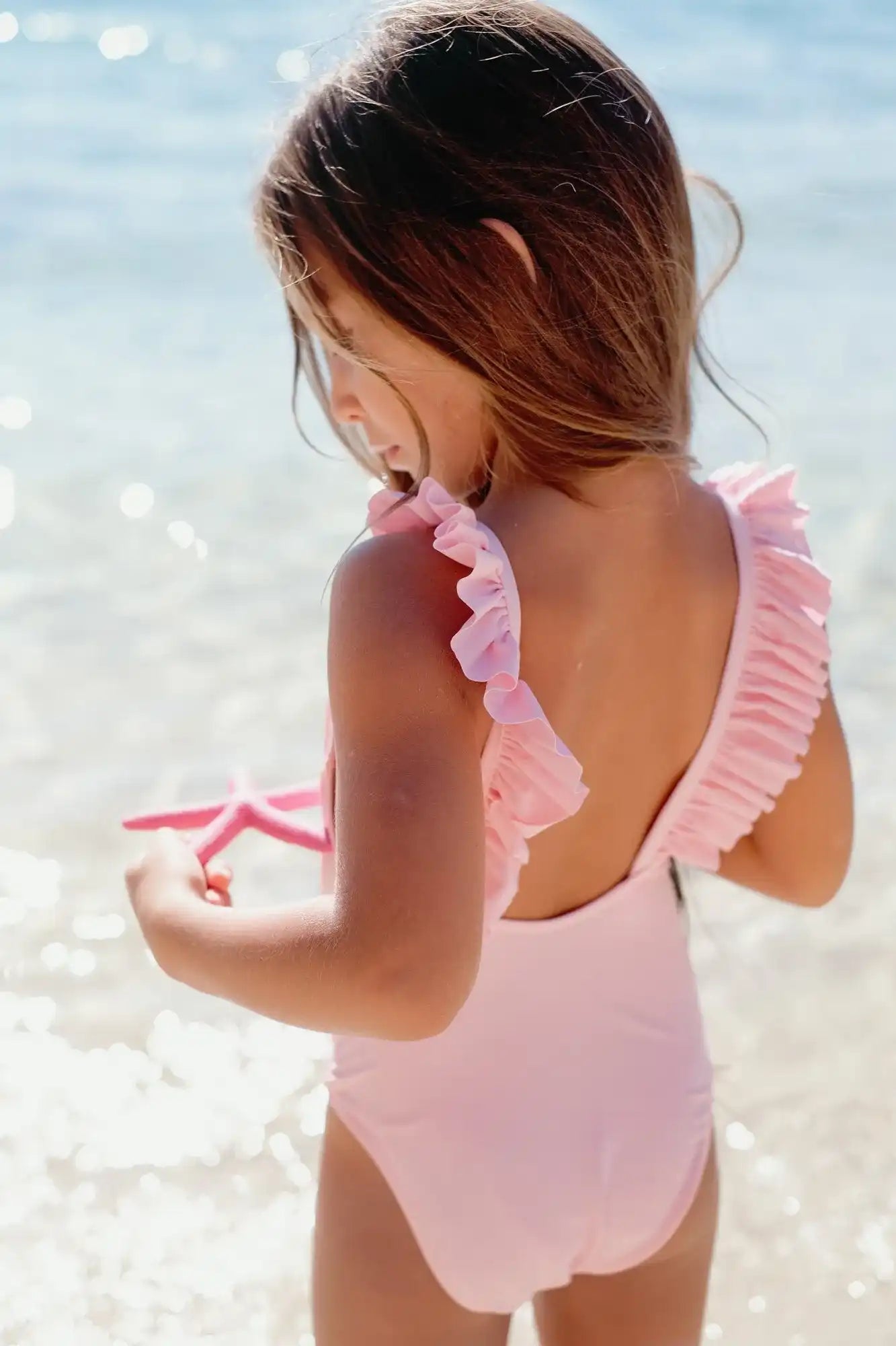 Girl in pale pink ruffled one-piece swimsuit by Lison Paris on the beach holding a starfish
