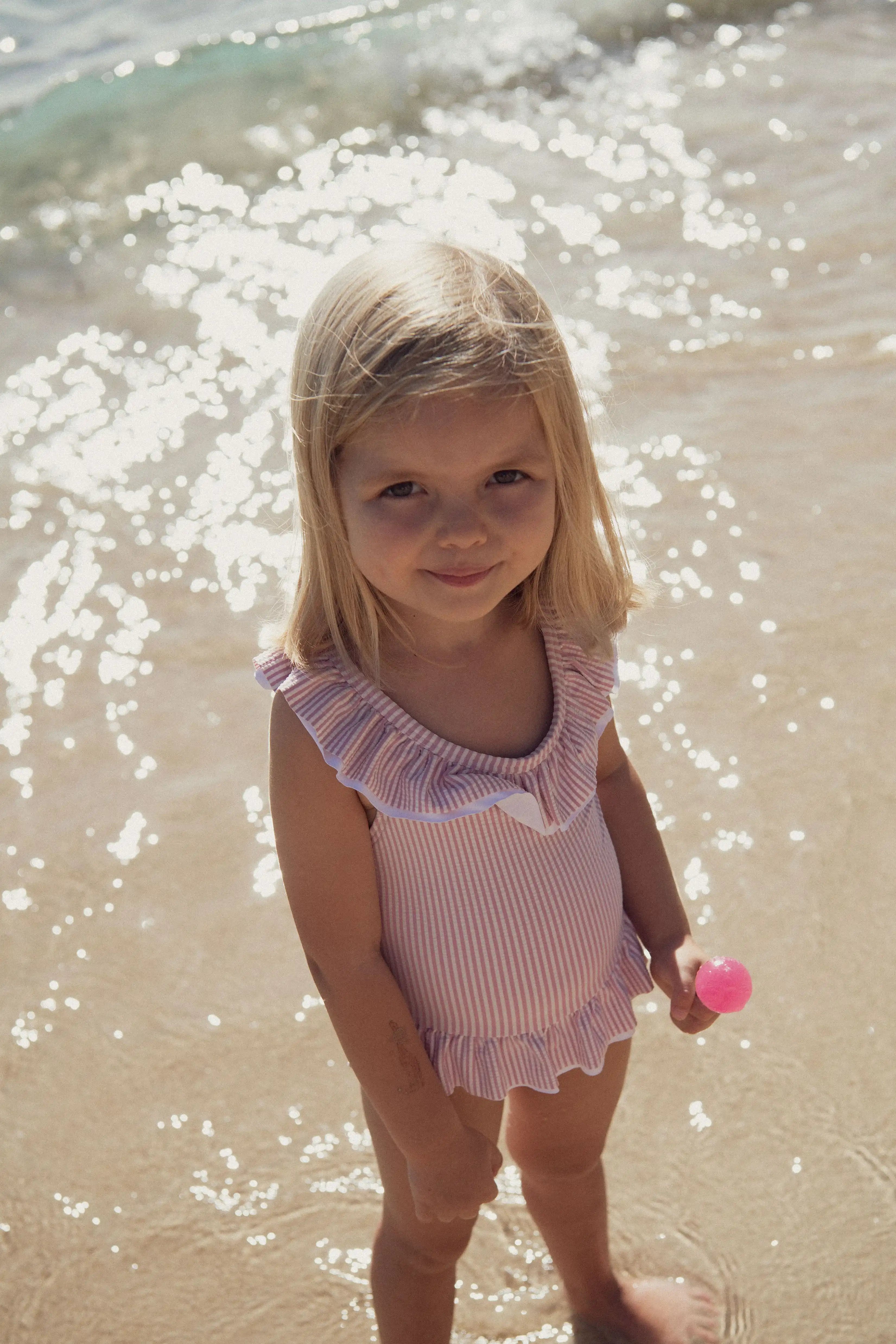 Young girl in Lison Paris lilac ruffled one-piece swimsuit on beach, sunlight on water
