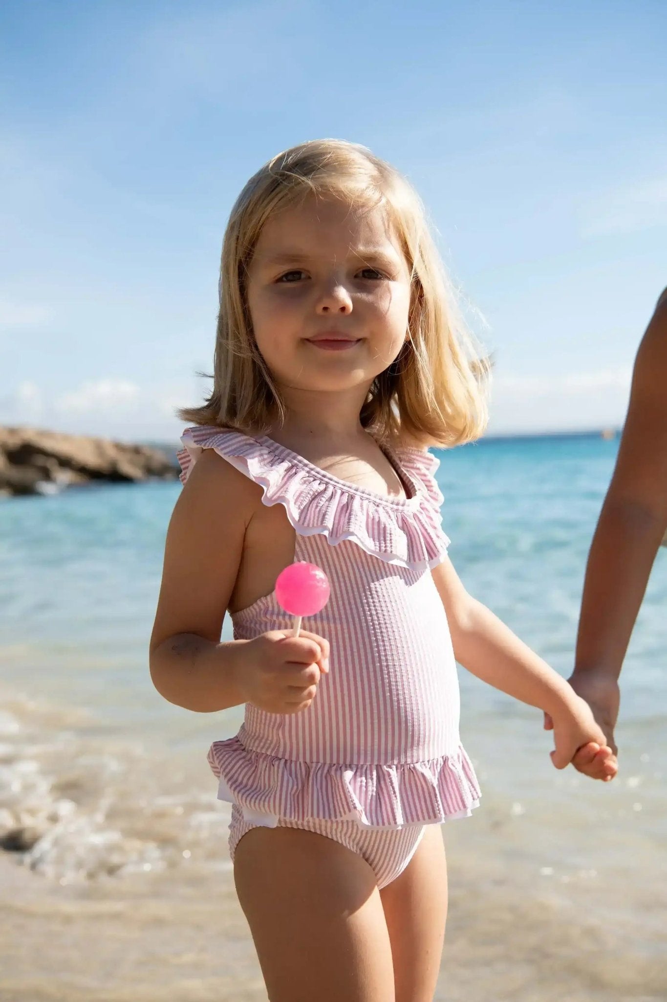 Girl in a lilac ruffled one-piece swimsuit holding a lollipop on a sunny beach