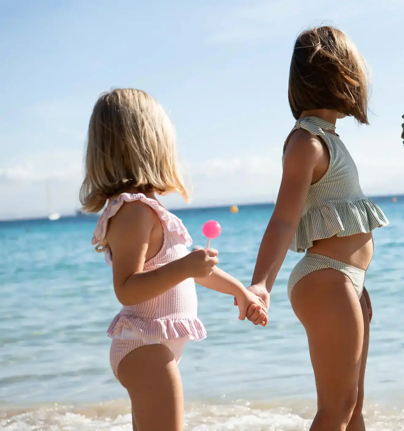 Two girls in ruffle swimsuits holding hands on a sunny beach, one with a pink lollipop.