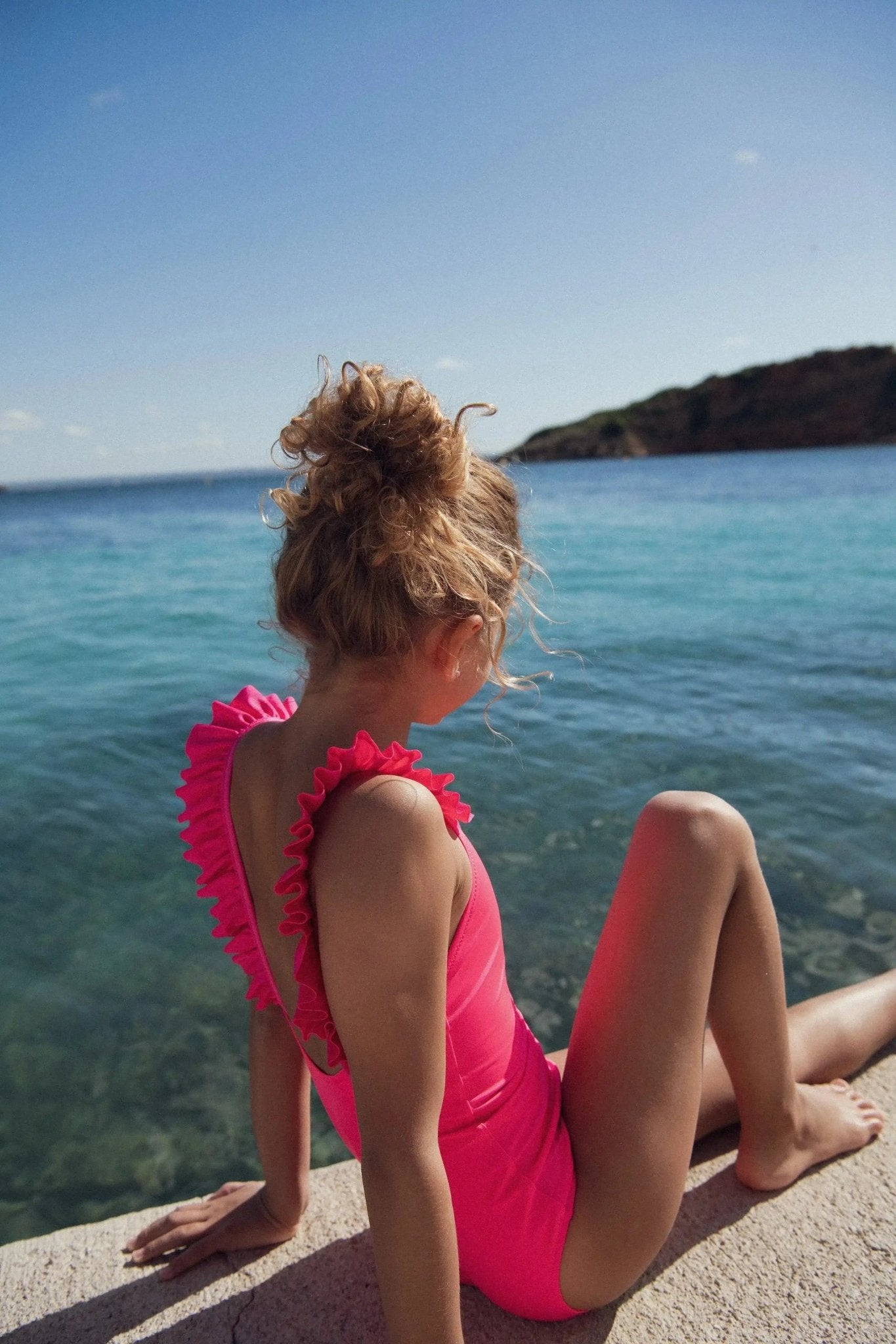 Girl in hot pink ruffled one-piece swimsuit sitting by the sea on a sunny day