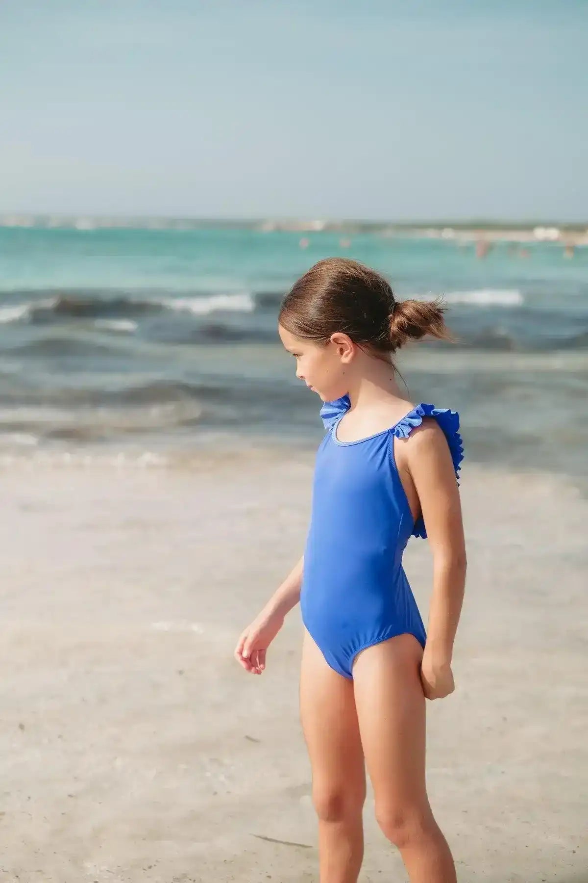 Girl on beach in blue ruffled one-piece swimsuit, ocean waves in background