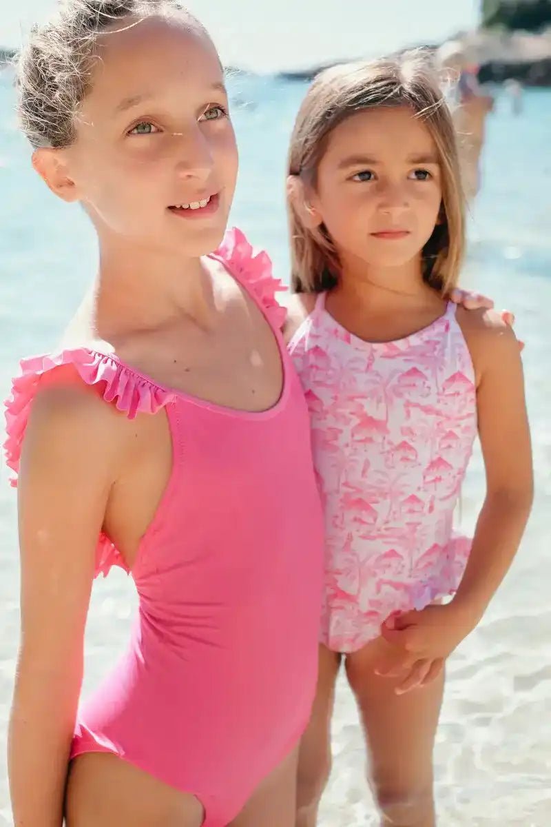 Two girls wearing pink one-piece swimsuits with ruffles standing by the seaside
