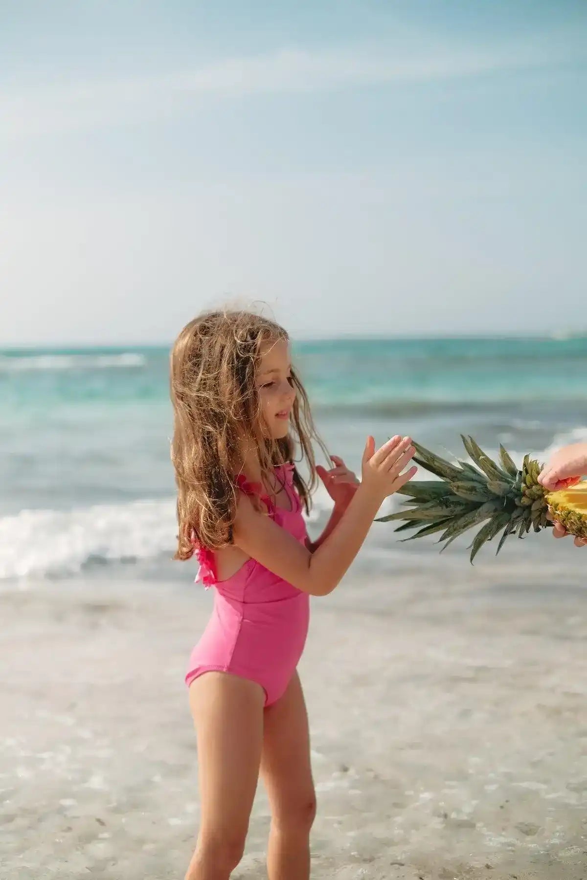 Girl in pink one-piece ruffled swimsuit on beach holding pineapple, UV-protective swimwear