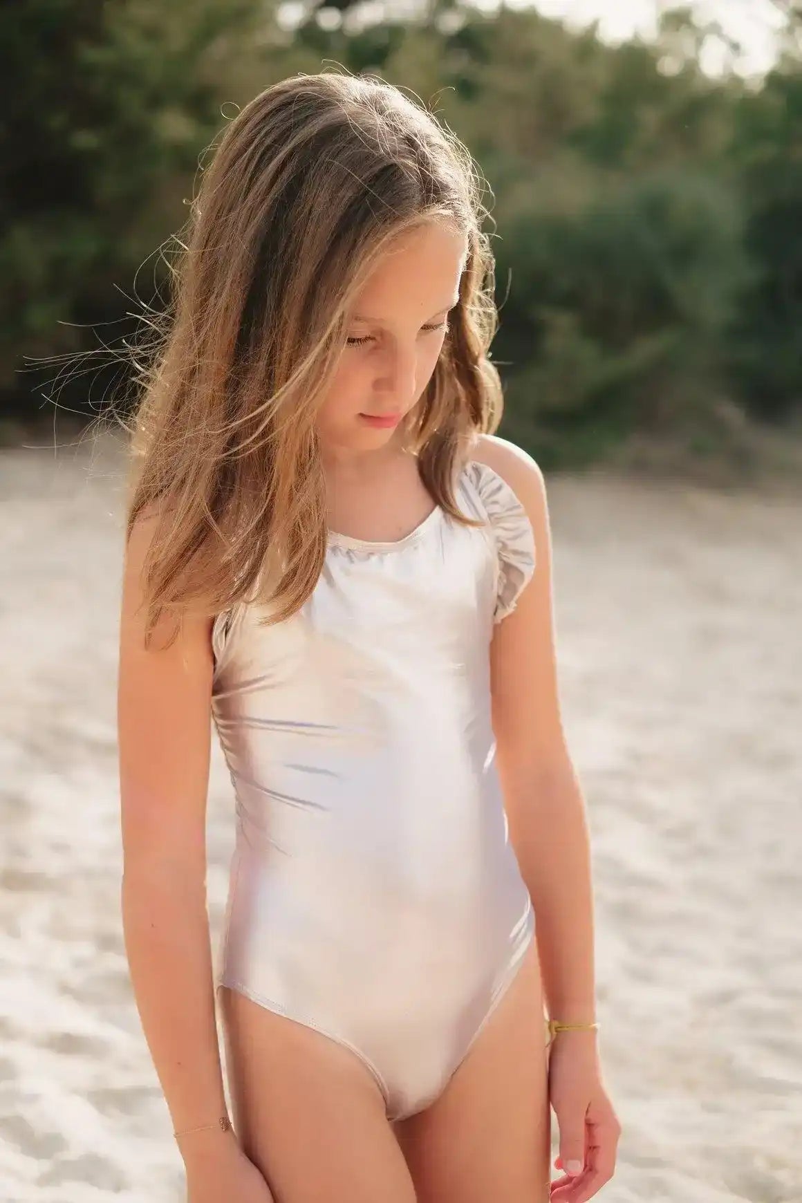 Girl in iridescent silver one-piece swimsuit on sandy beach with green trees