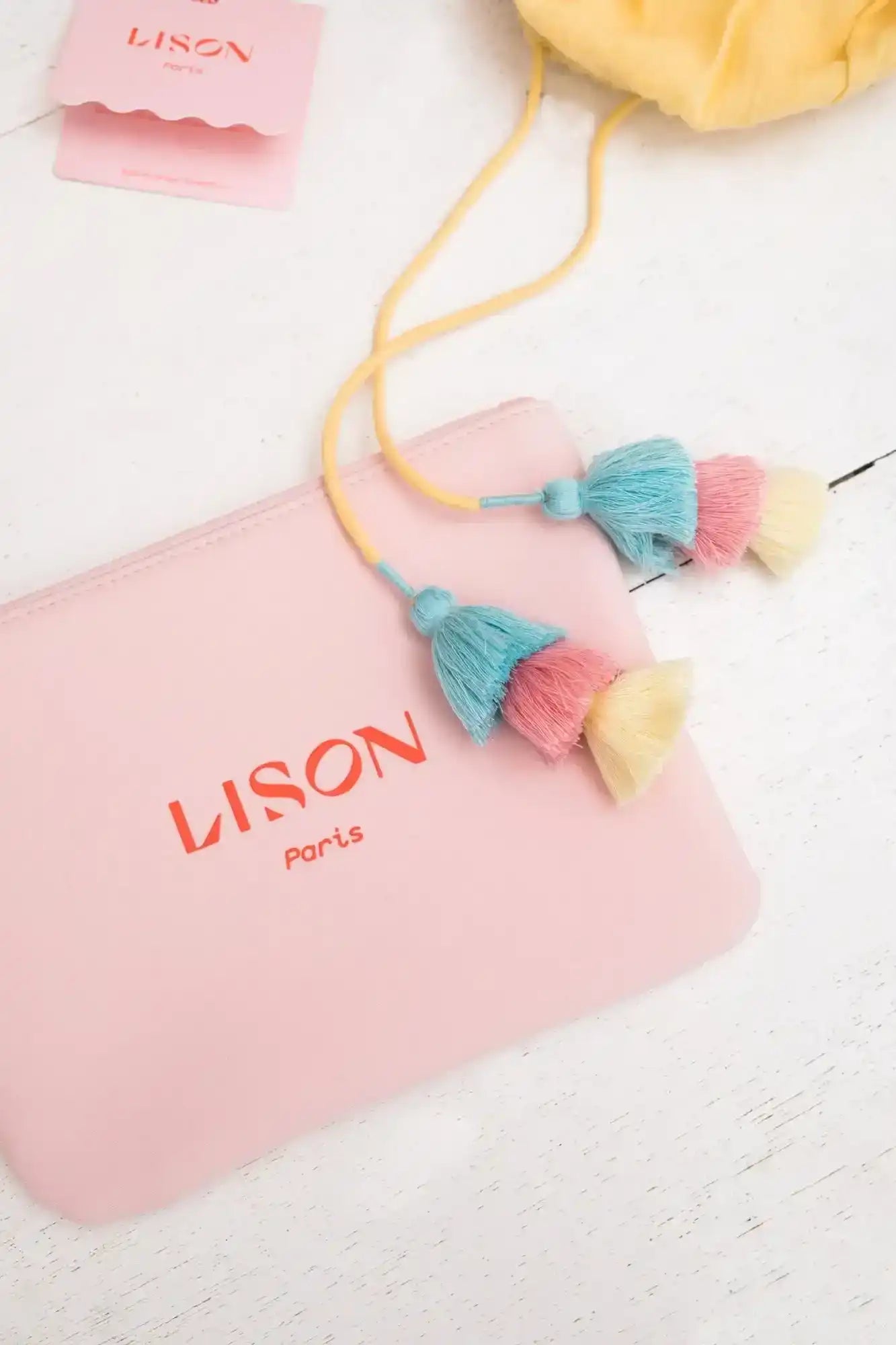 Pink Lison Paris pouch with yellow drawstring and pastel tassels on white surface