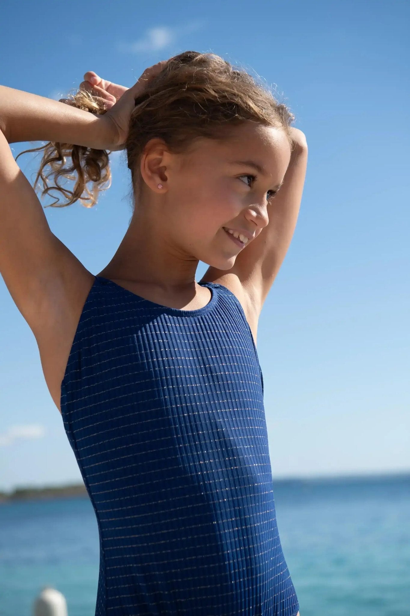 Smiling girl in navy blue one-piece swimsuit with gold stripes by the sea on a sunny day