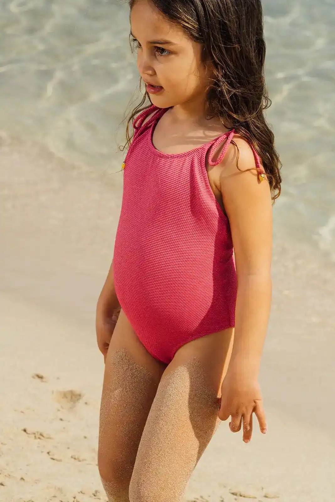 Girl in a pink textured one-piece swimsuit standing on sandy beach by the water