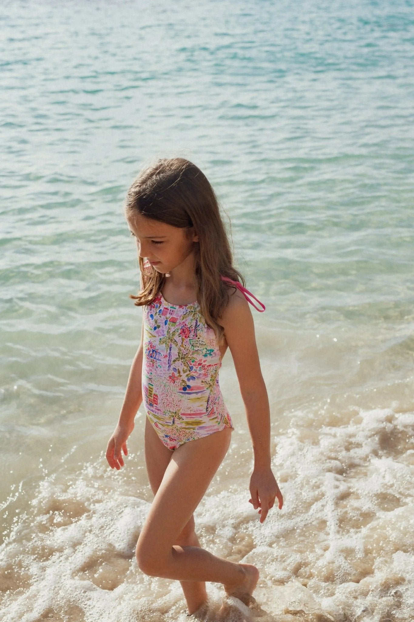 Girl in pink floral one-piece swimsuit walking on beach by the sea