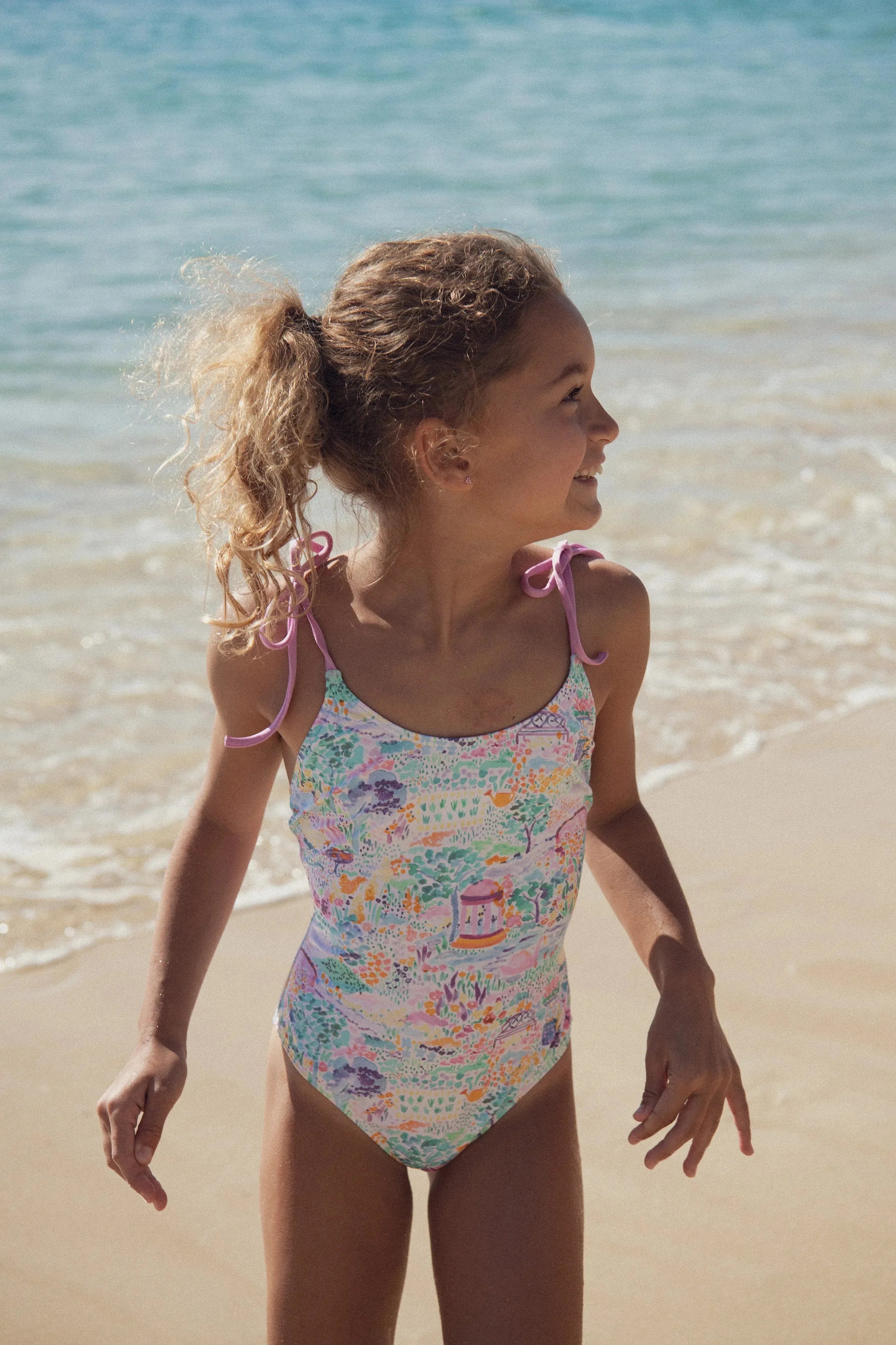 Girl wearing Lison Paris pink floral one-piece swimsuit on a sunny beach