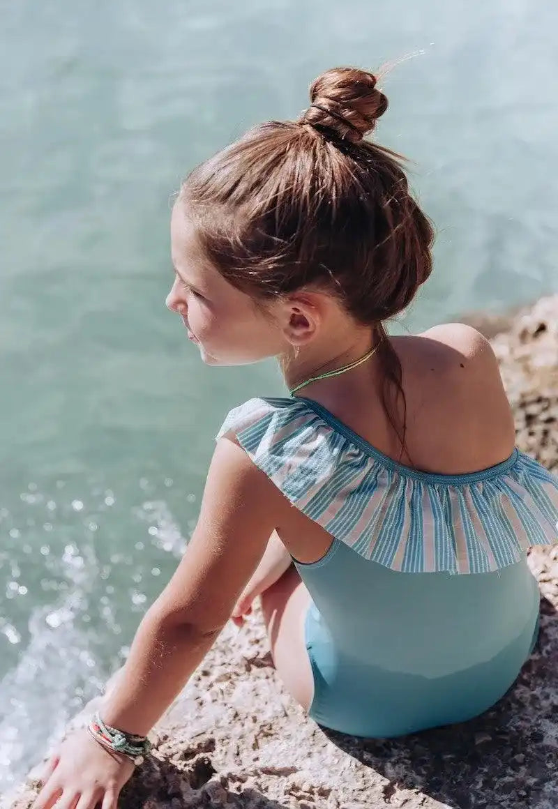 Girl in Lison Paris blue one-piece ruffled swimsuit by the water, enjoying sunny beachwear