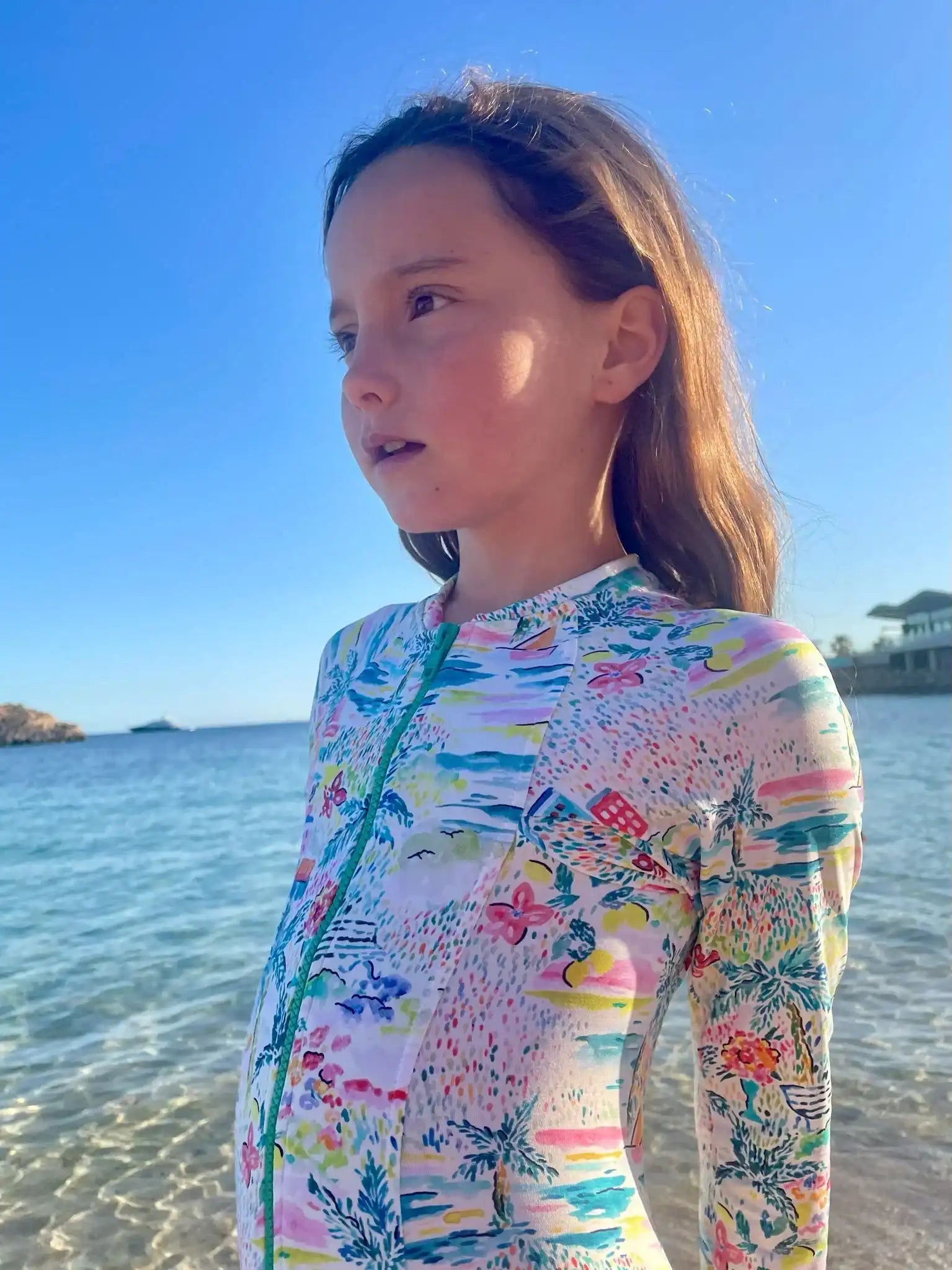 Girl wearing Lison Paris long-sleeve floral swimsuit on a sunny beach, clear blue sky