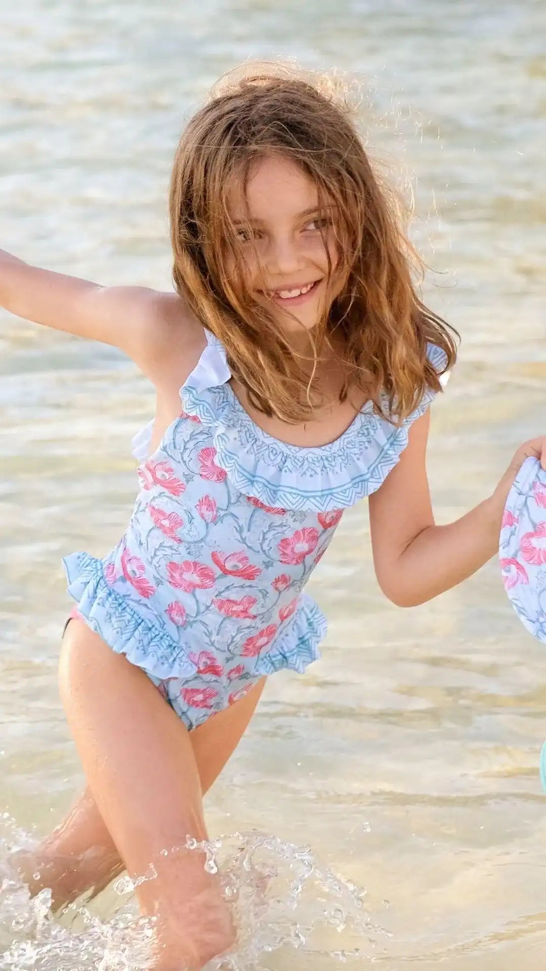 Girl in a blue floral Lison Paris one-piece swimsuit with ruffles playing in shallow water