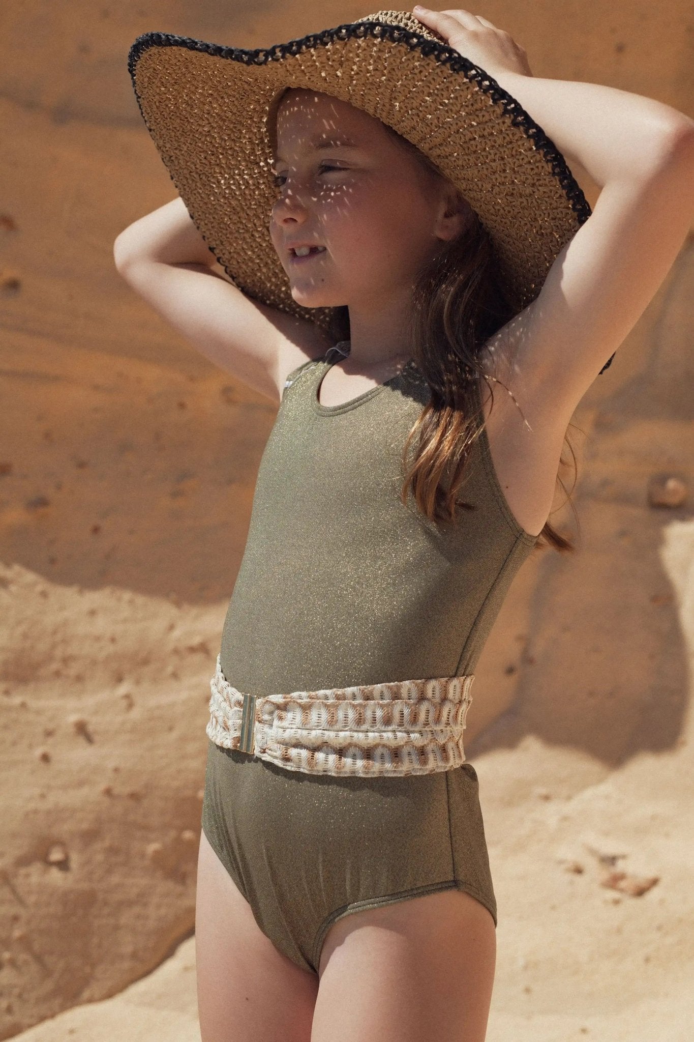Girl in khaki one-piece swimsuit with decorative belt and straw hat on a sandy beach