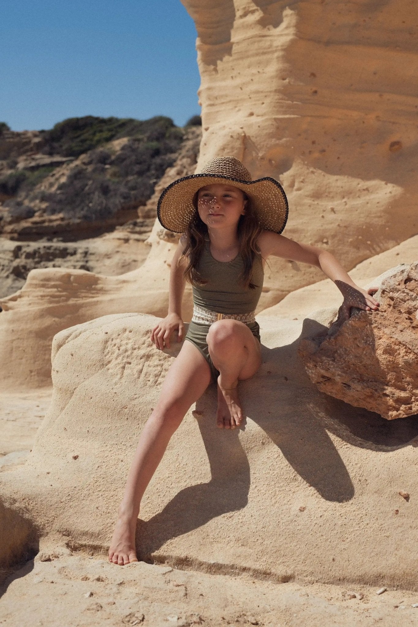 Girl in khaki one-piece swimsuit and straw hat sitting on rocks at the beach