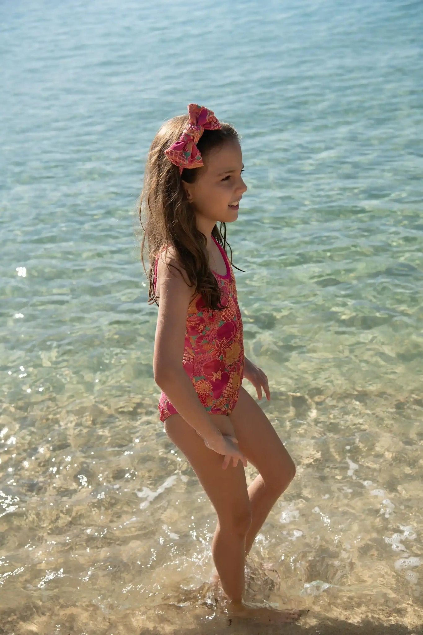Girl in pink floral one-piece swimsuit and matching headband standing in shallow sea water
