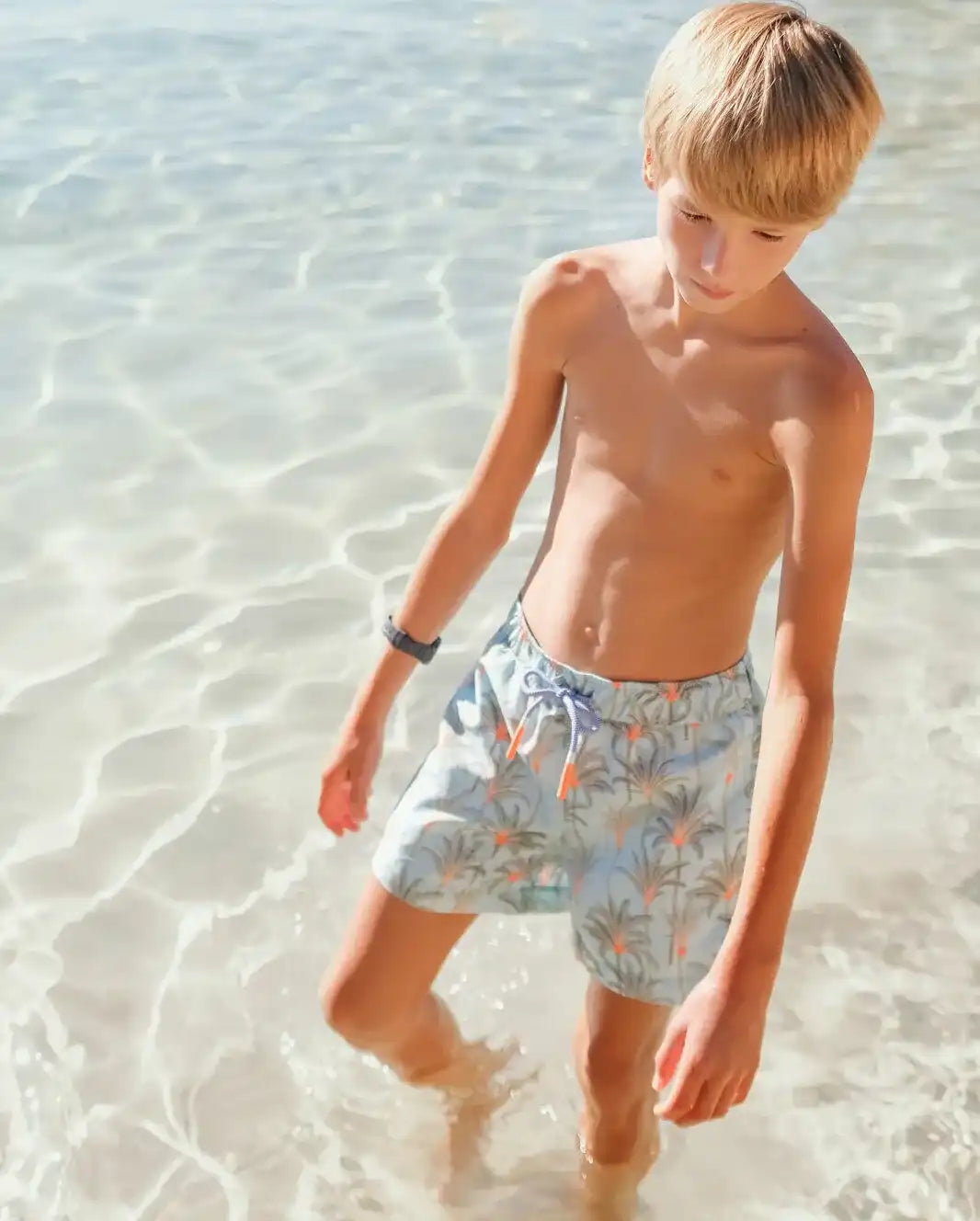 Boy wearing Lison Paris aqua palm swim shorts walking in clear beach water