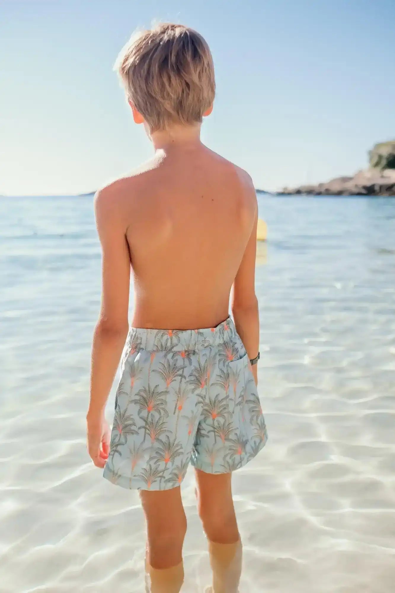 Boy in aqua palm tree print swim shorts standing in clear shallow beach water