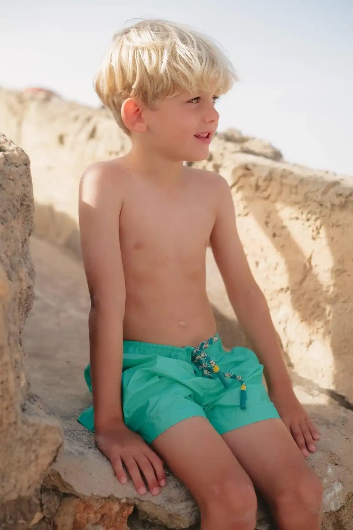 Boy in aqua swim shorts sitting on rocks, Lison Paris beachwear collection, sunny day