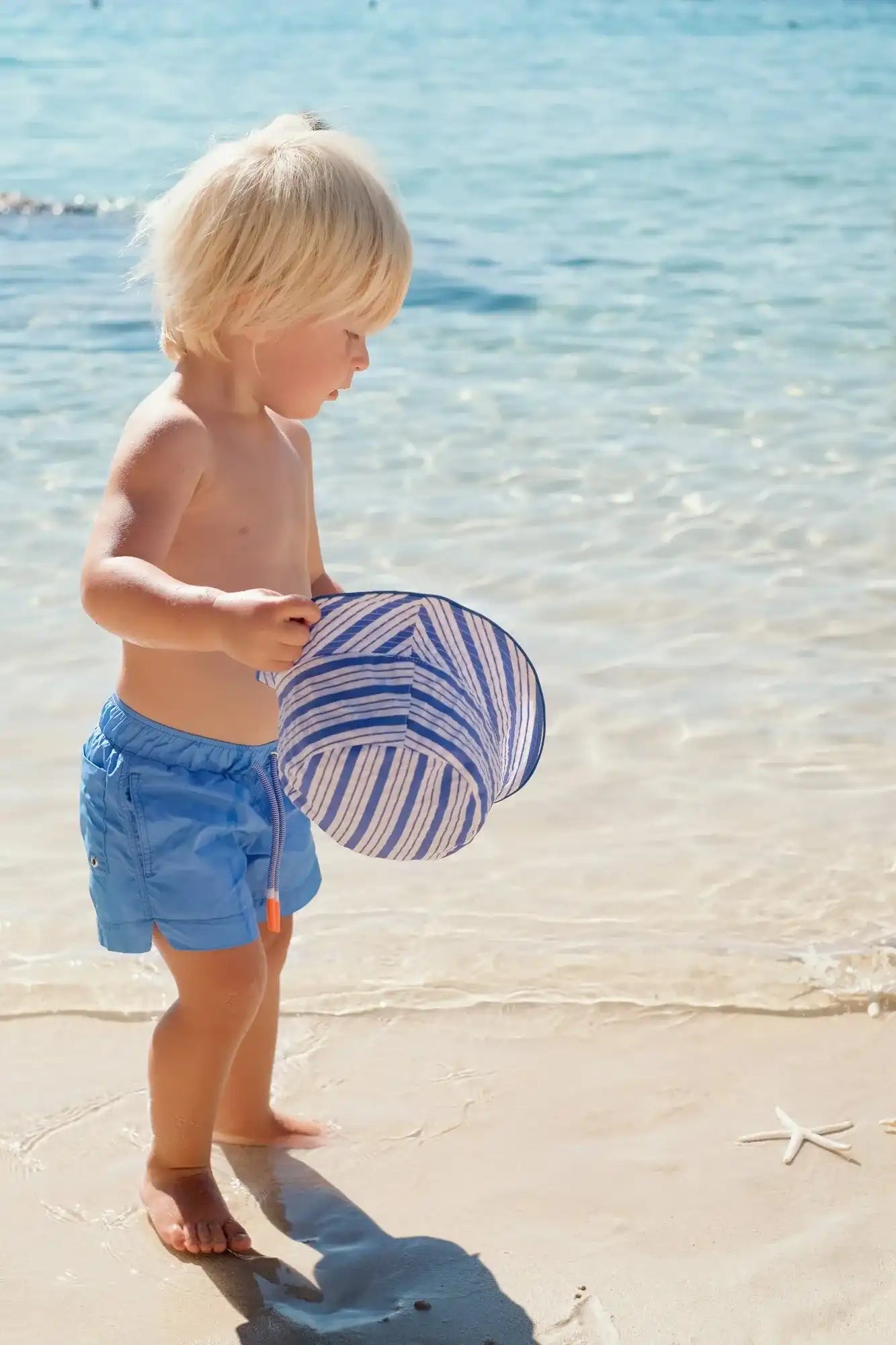 Young boy in blue swim shorts holding striped hat on sunny beach, Lison Paris swimwear