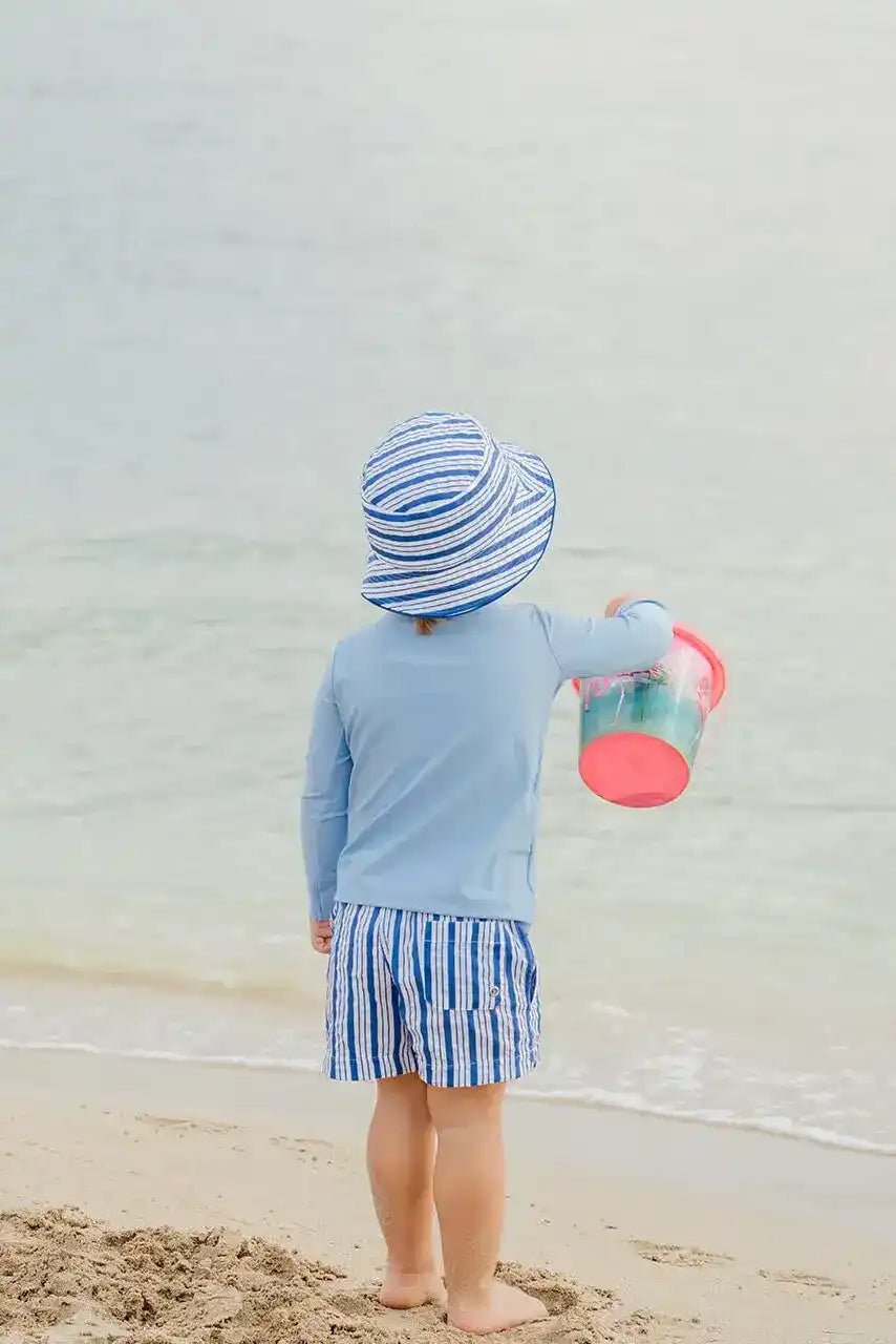 Boy in blue striped swim shorts and sun hat holding bucket on sandy beach