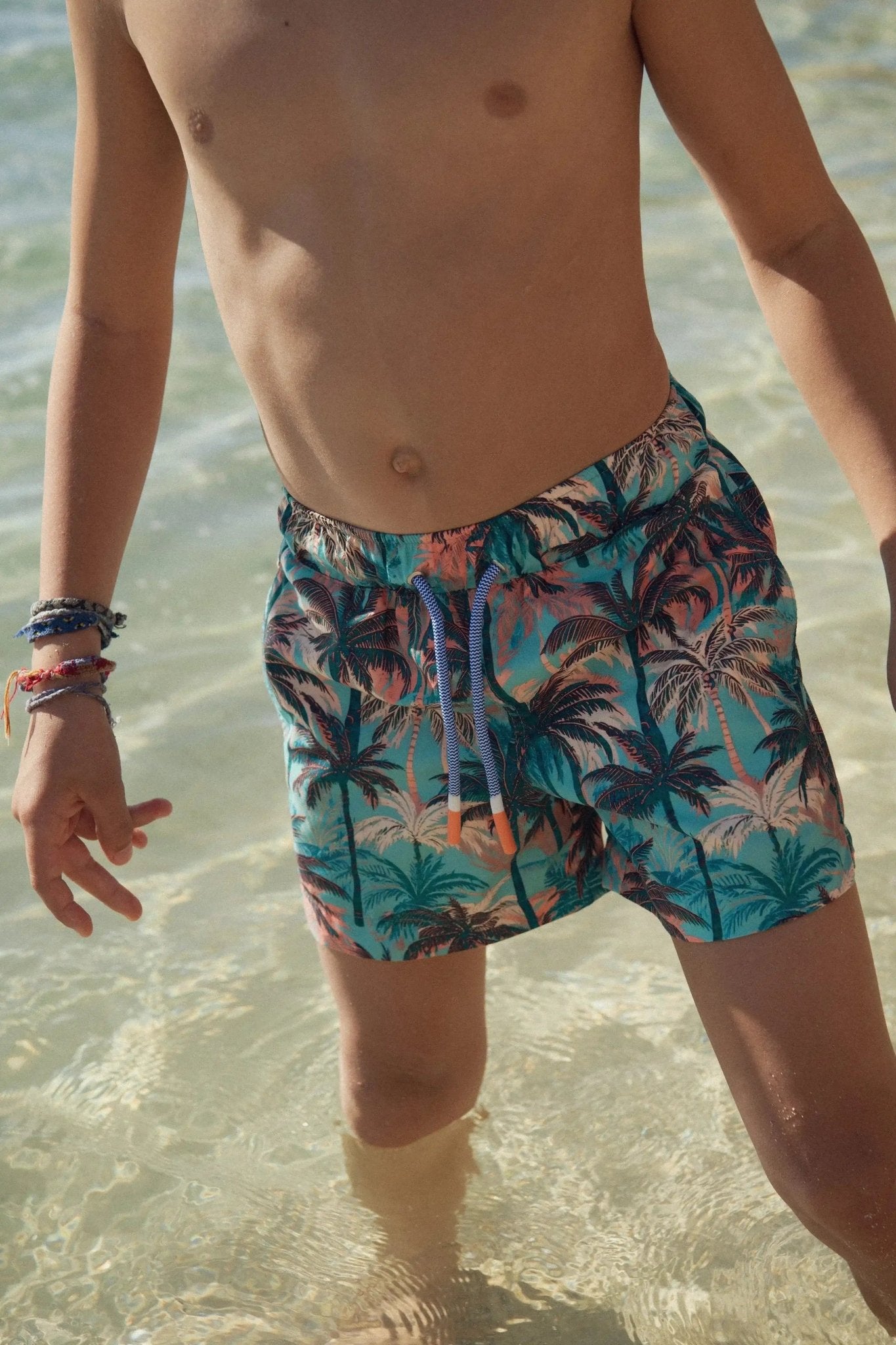 Boy wearing blue and orange palm tree swim shorts standing in shallow beach water