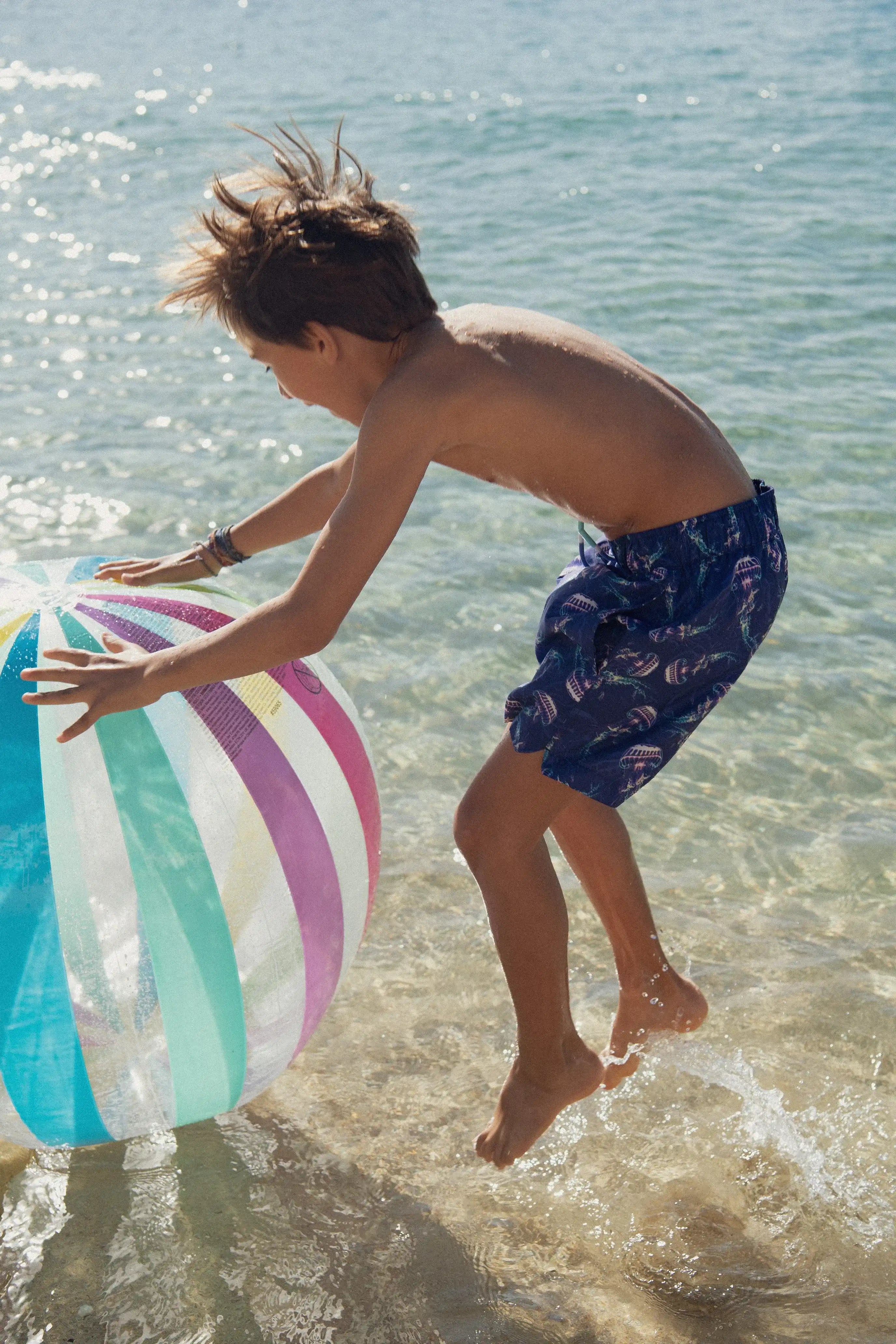 Boy in blue jellyfish print swim shorts playing with beach ball in shallow sea, Lison Paris