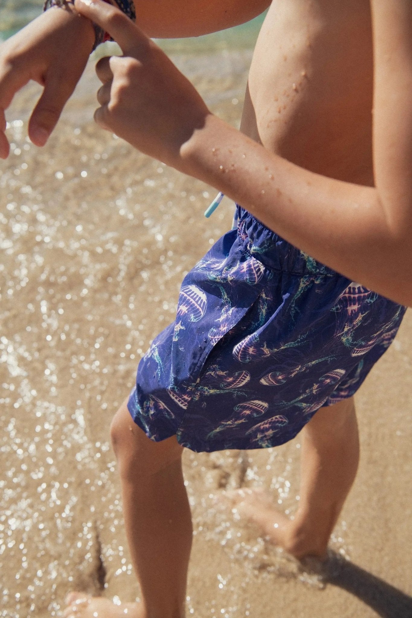 Boy wearing blue jellyfish print swim shorts at the beach, standing in shallow water