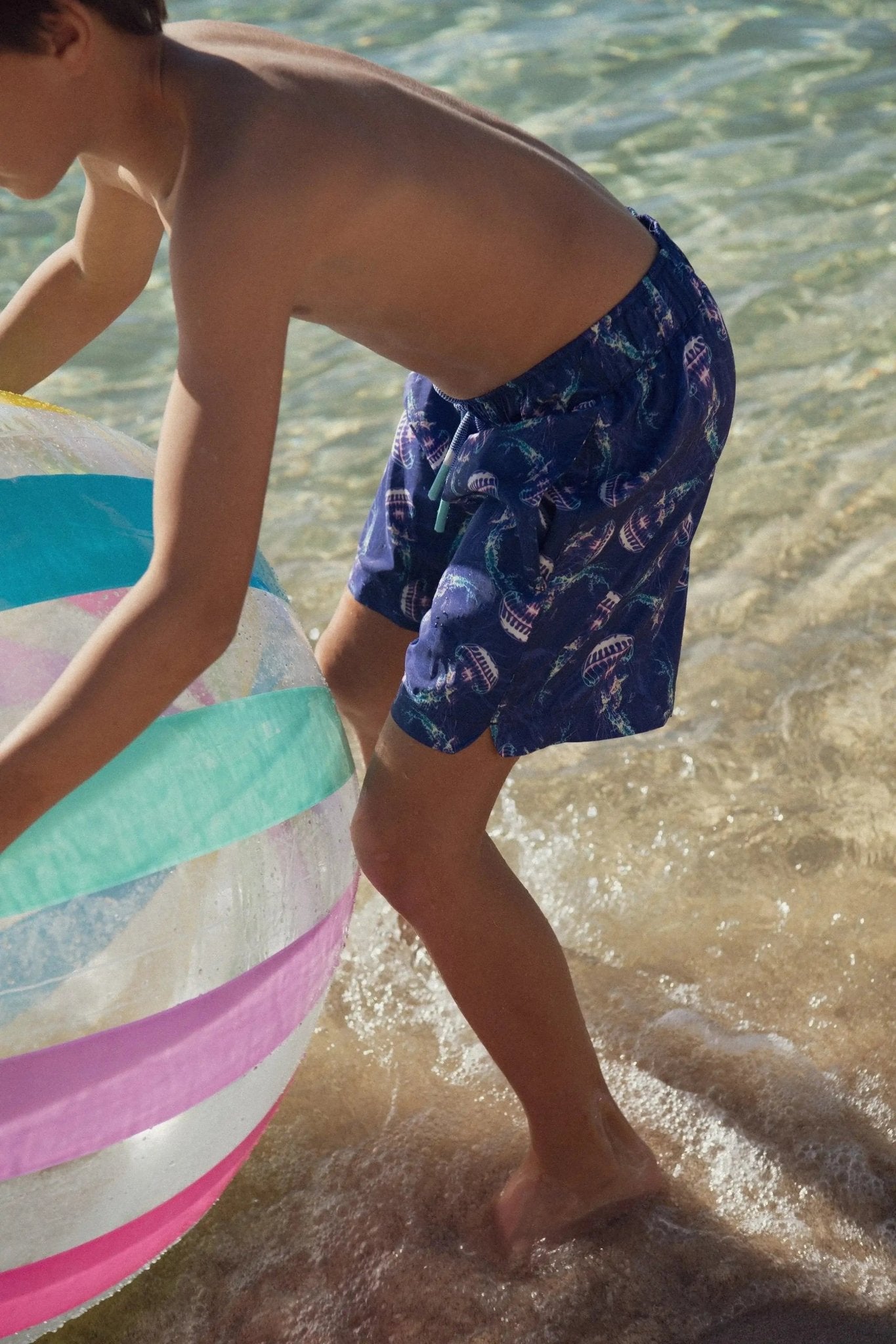 Boy in blue jellyfish print swim shorts playing with beach ball at the water's edge
